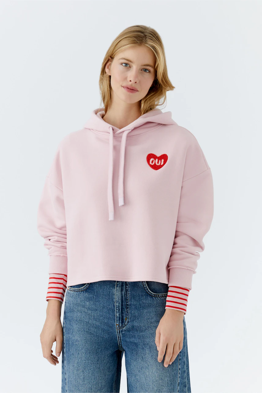 Casual women's hoodie: Young woman in a pink cropped pullover with red heart detail and striped sleeves.