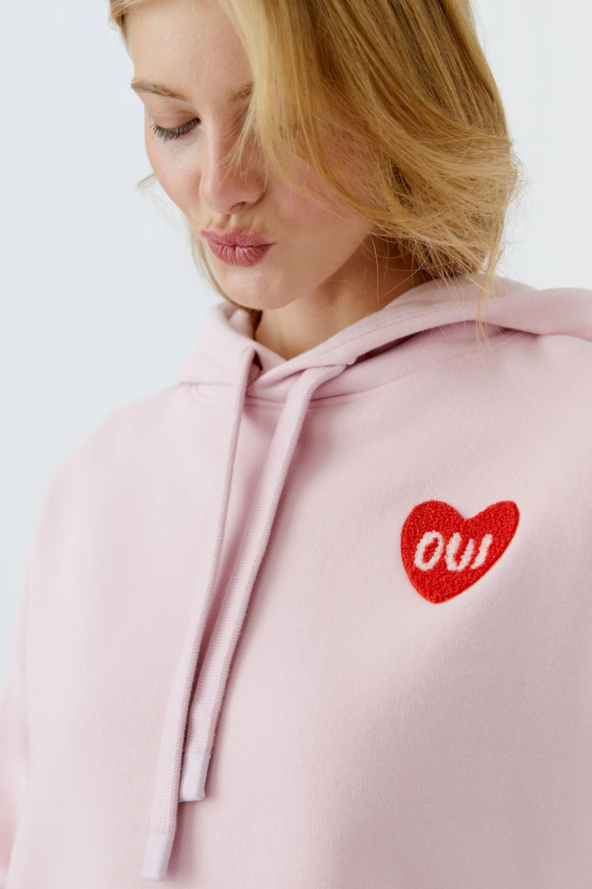 Casual women's hoodie: A woman in a light pink pullover with a red heart and 'oui' logo.