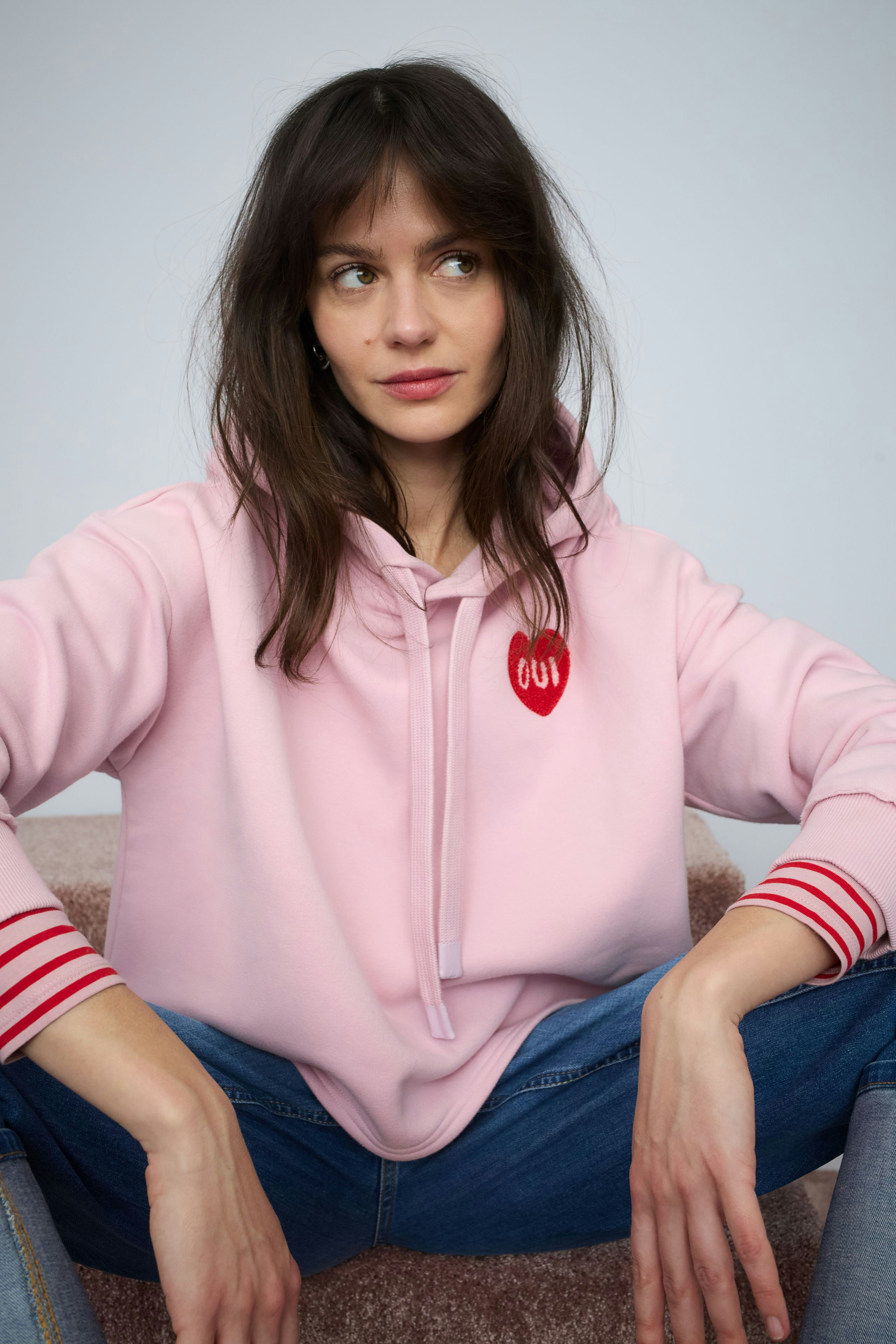 Bequemer Damen-Hoodie: Frau in rosa Kapuzenpullover mit roten Streifen und Herz-Logo sitzt entspannt.