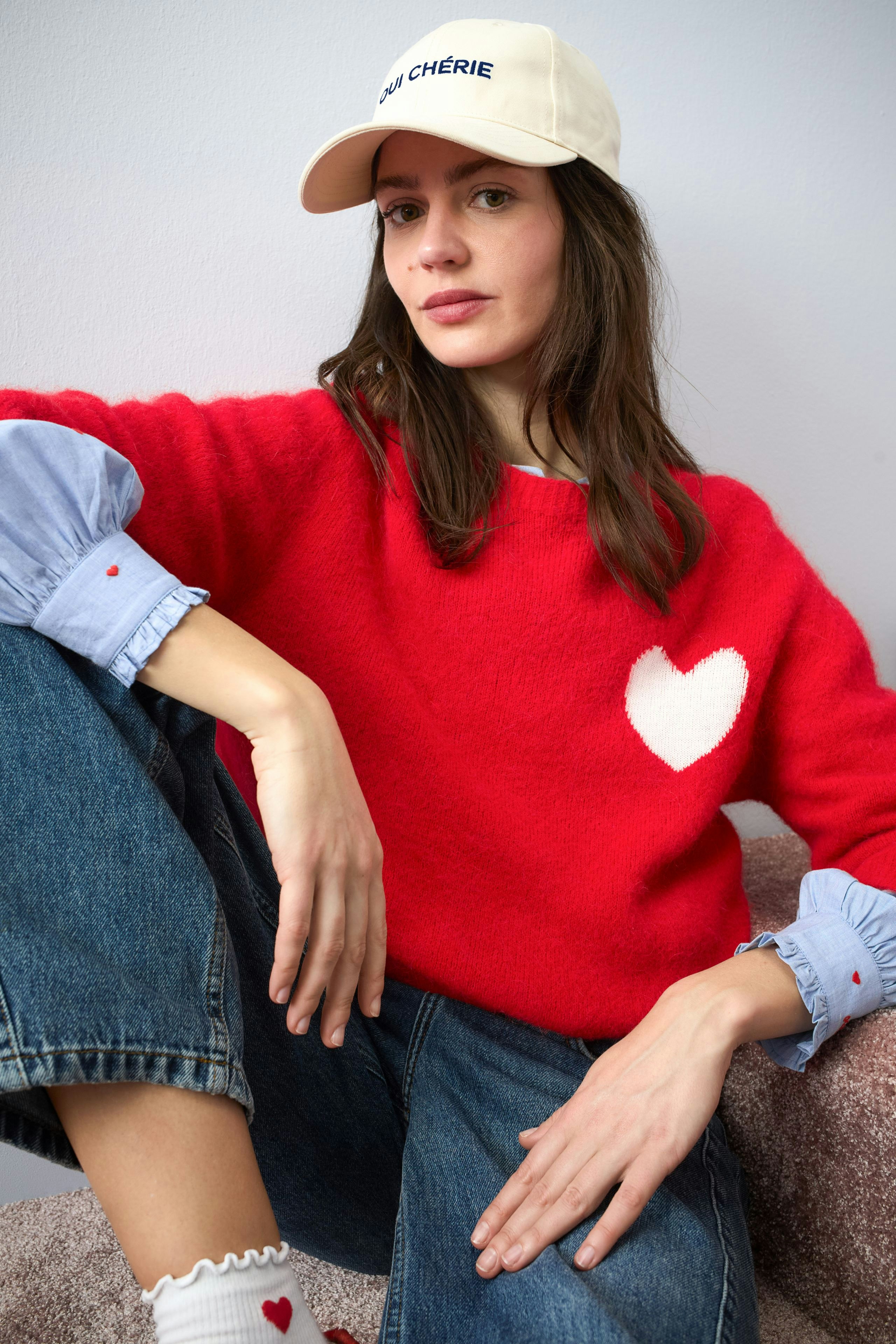 Casual women's hat: A woman wearing a cream cap with 'Oui Chérie' and a cozy red pullover with a heart.