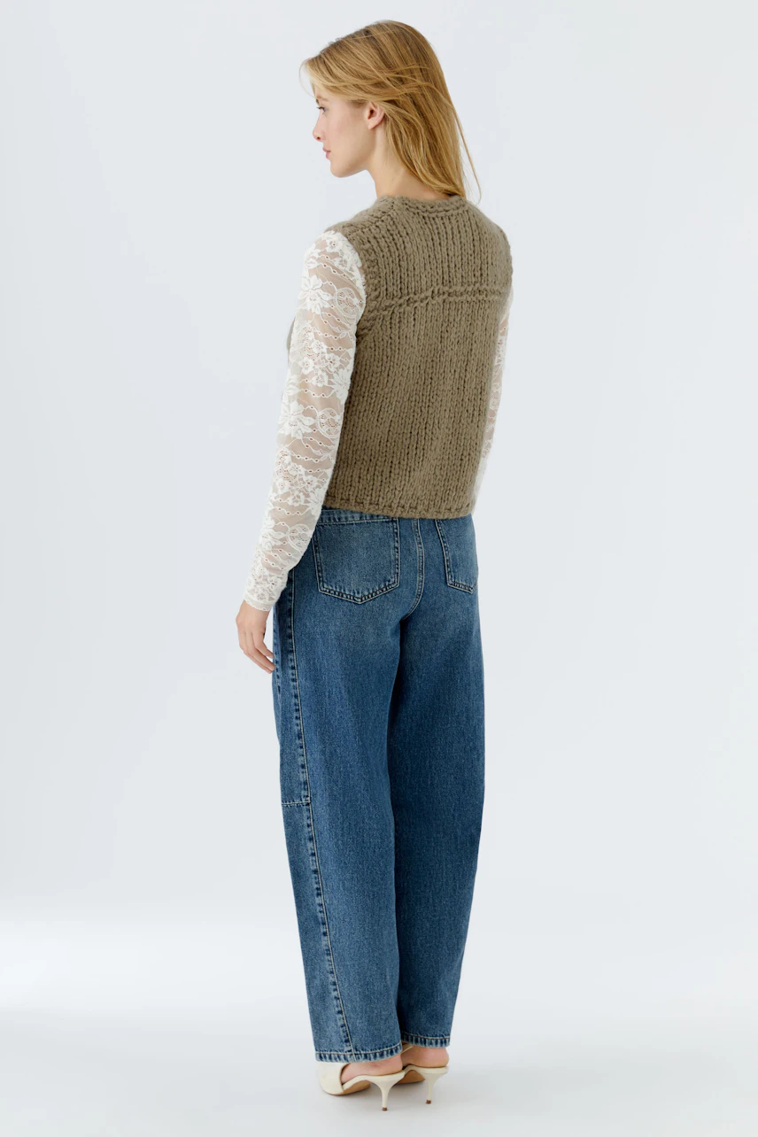 Damen-Strickweste: Frau mit langärmligem Oberteil und lässiger Jeans trägt eine olivgrüne Strickweste.