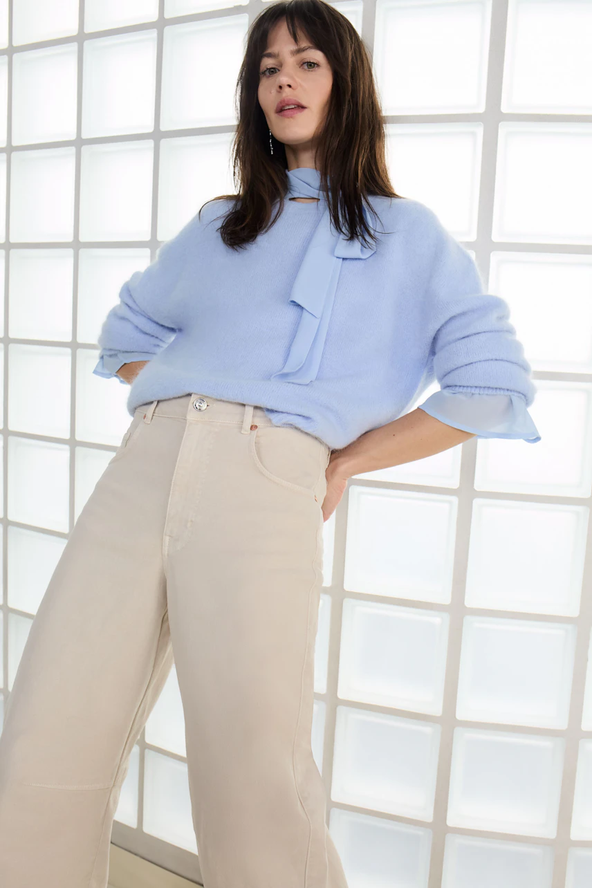 Damen-Bluse: Frau in blauer Bluse mit Schleife und beigen Hosen, lässiger Look vor hellen Wänden.