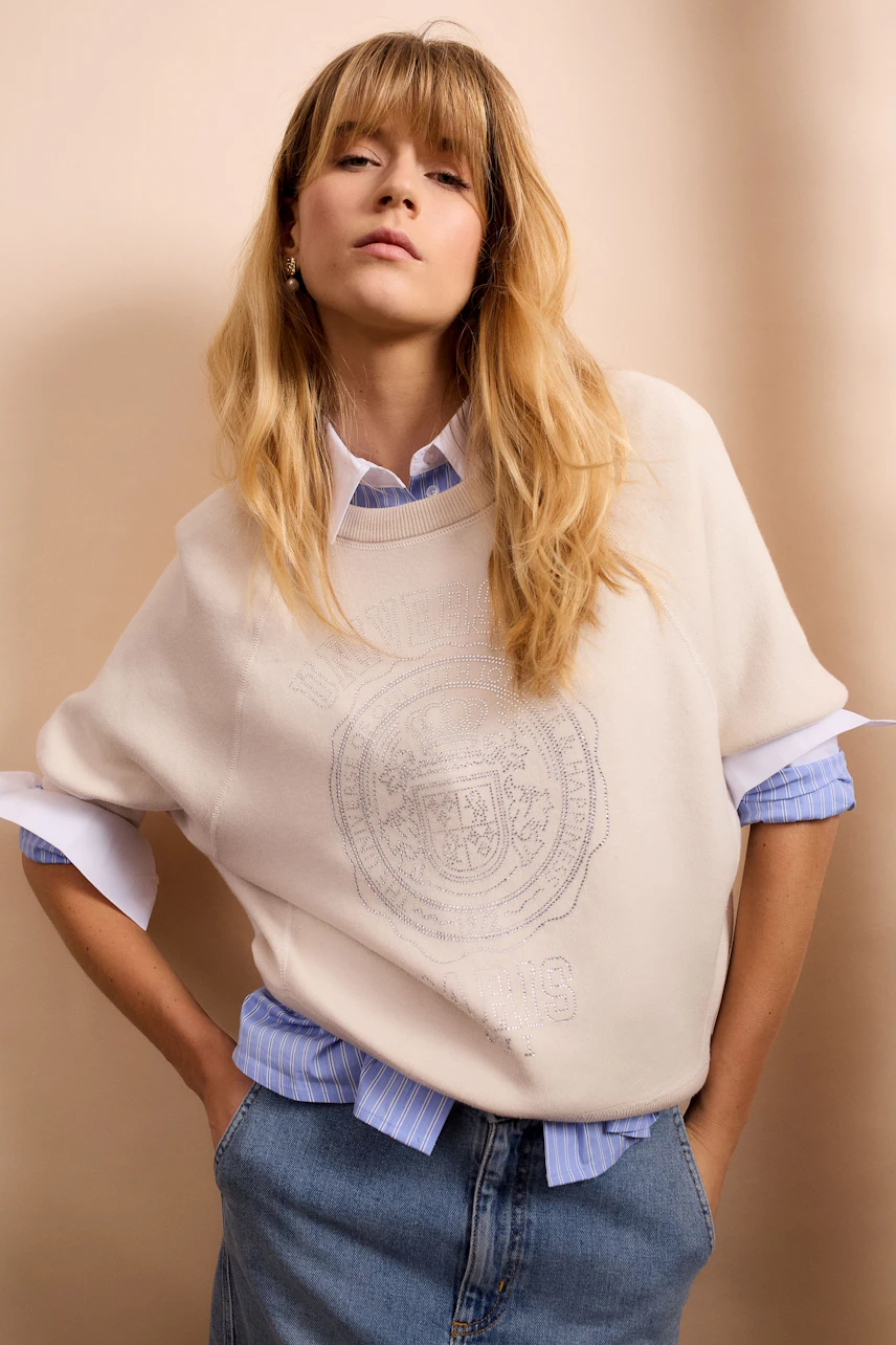 Casual women's pullover: Young woman in a light pullover with a logo, layered over a blue shirt.