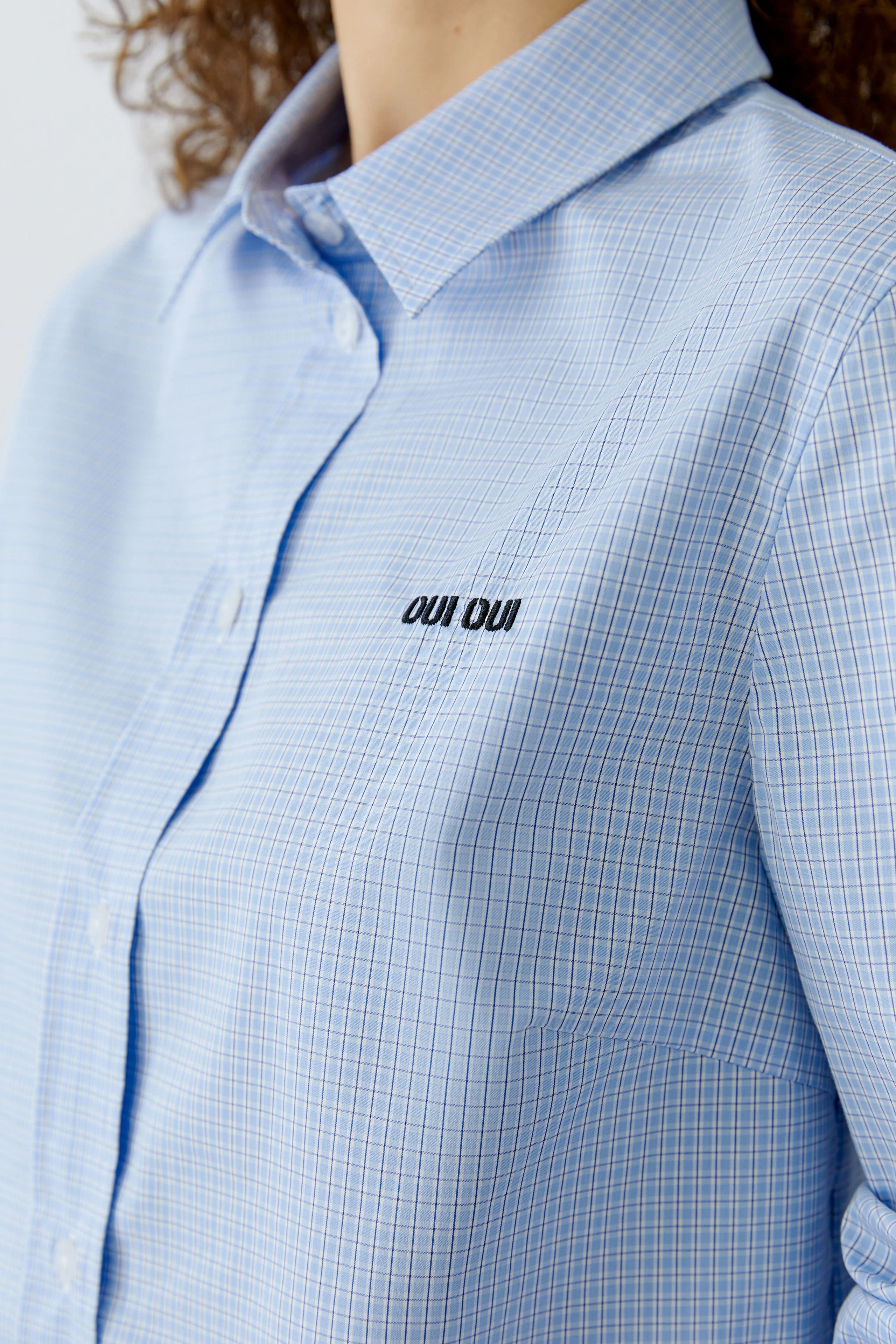 Casual women's shirt blouse: Close-up of a woman wearing a light blue checkered shirt with logo.