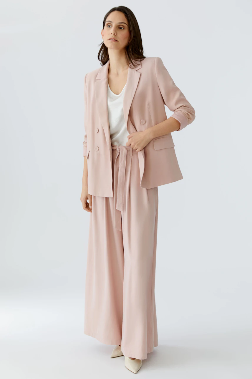 Damen-Bundfaltenhose in zartem Rosa, kombiniert mit einem lässigen Blazer und einem weißen Top.