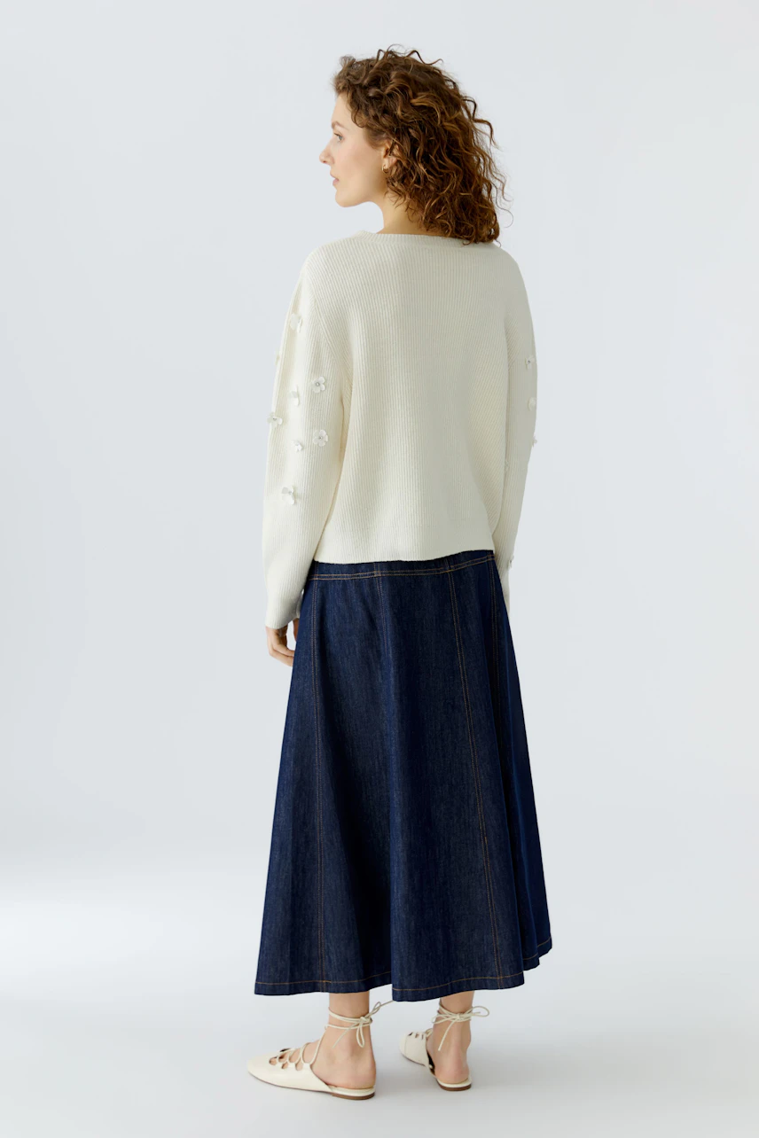 Damen-Pullover: Frau mit lockigem Haar trägt einen hellen Pullover mit Blumenstickerei und Jeansrock.