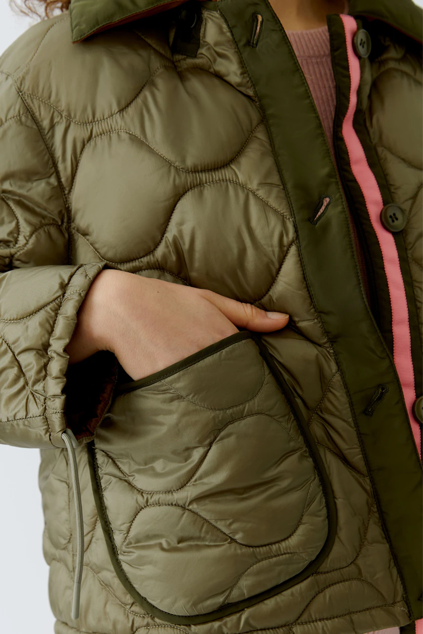 Wendejacke: Frau trägt eine olivgrüne, gesteppte Jacke mit pinken Akzenten und praktischen Taschen.