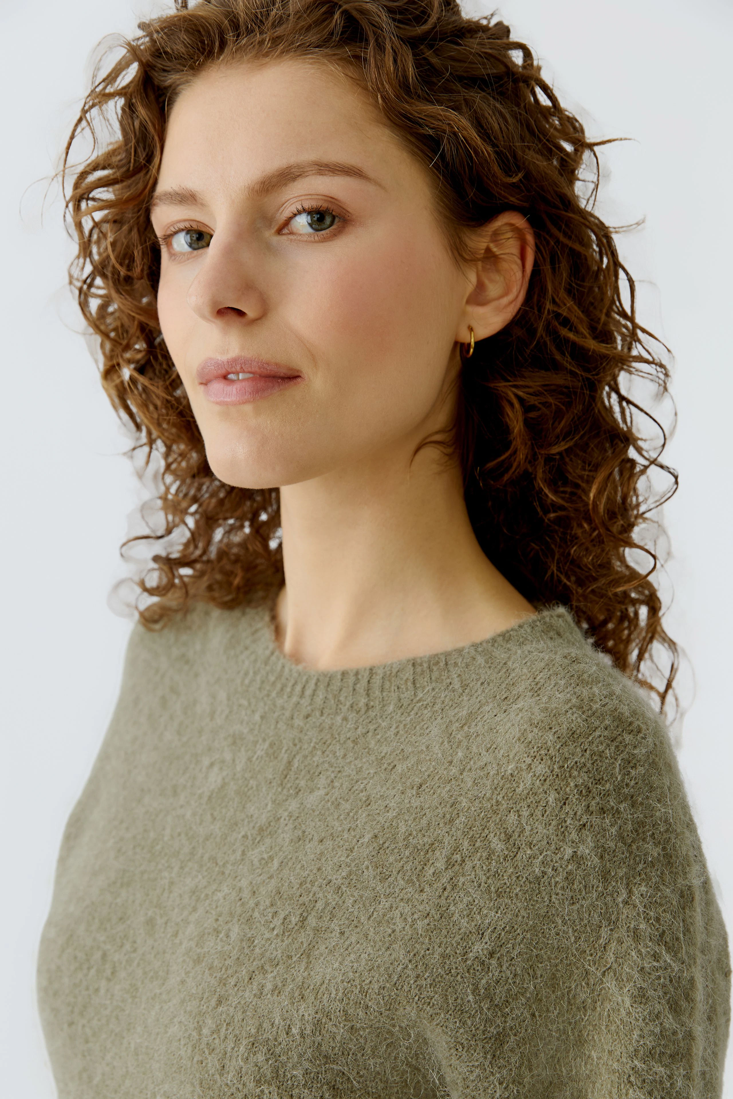 Damen-Pullover: Frau mit lockigem Haar trägt einen weichen, olivgrünen Strickpullover.