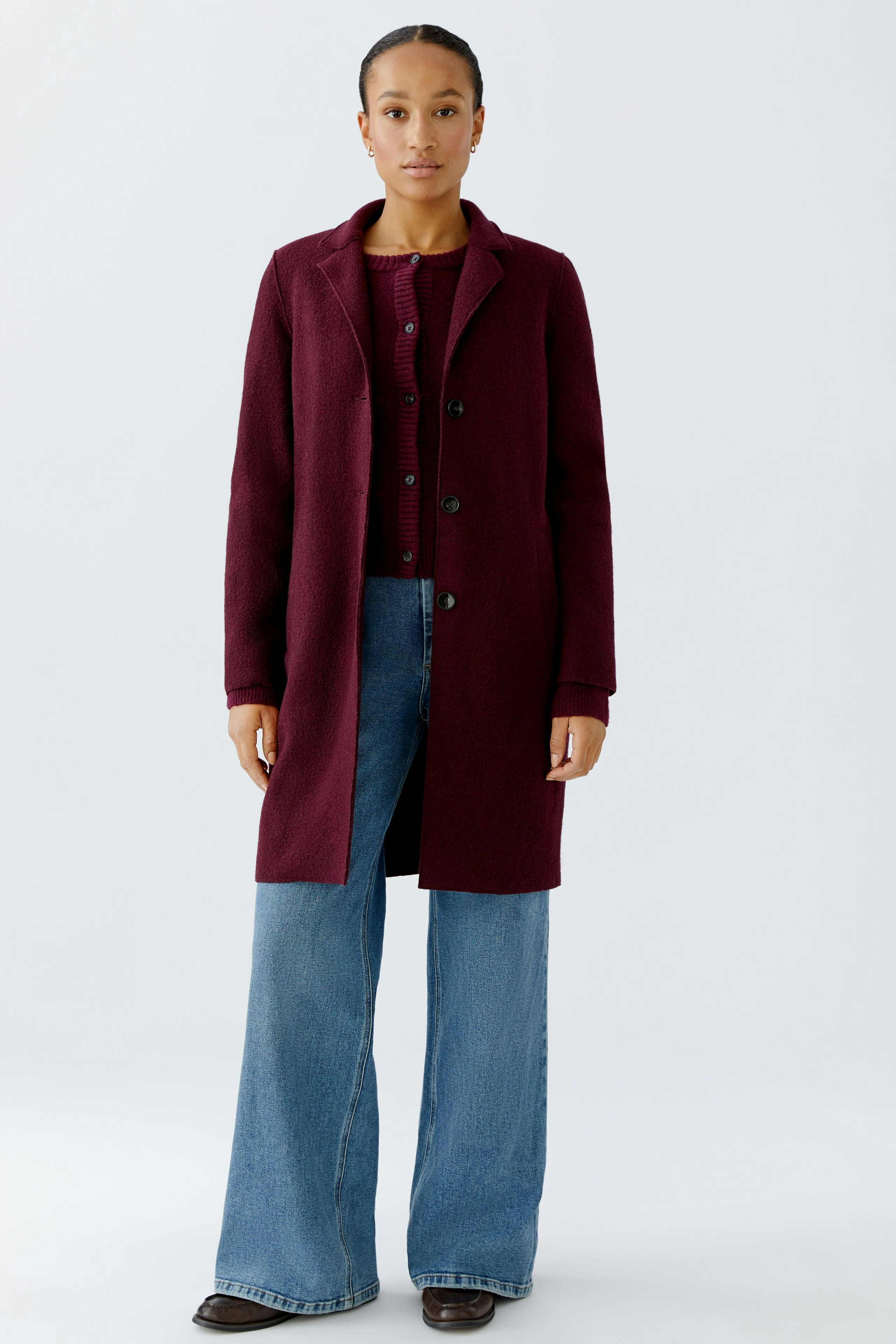 Modern women's wool coat: A woman in a burgundy coat over a matching cardigan and wide-leg jeans.