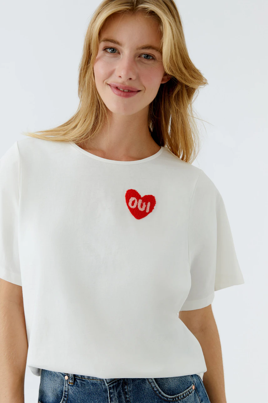 Casual women's T-shirt: Young woman in a white tee with a red heart design, smiling confidently.
