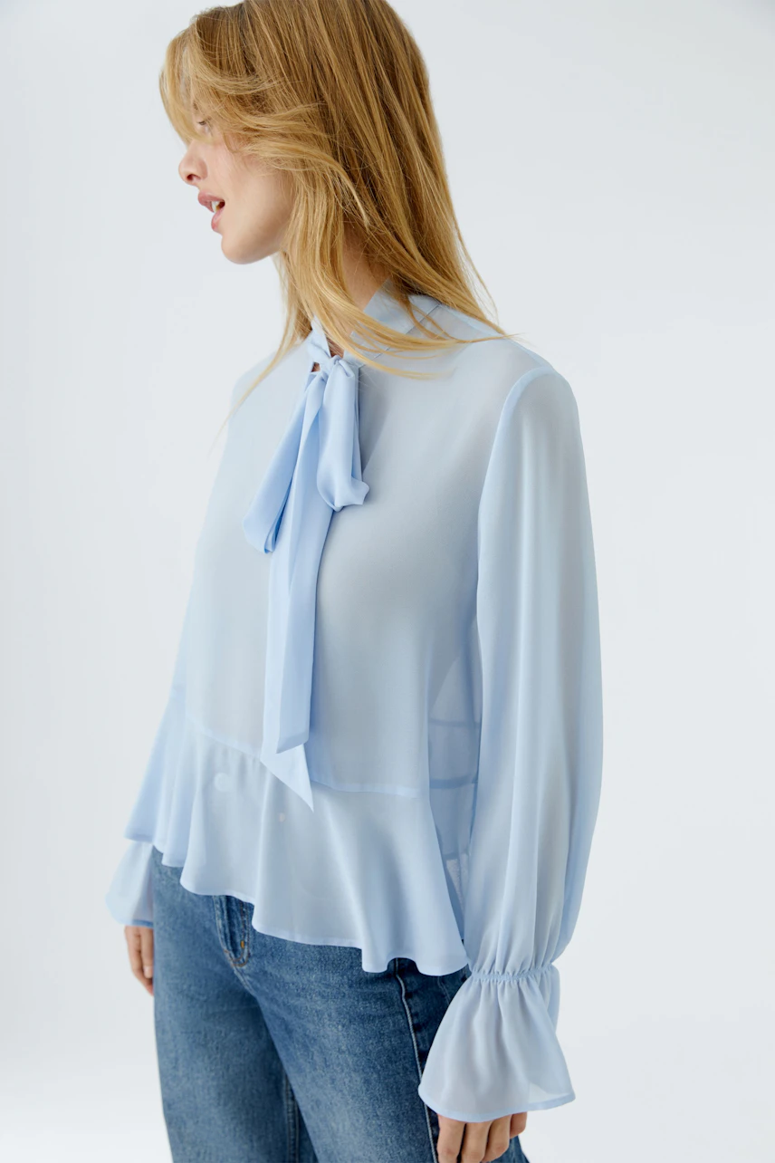 Leichte Damen-Bluse in Hellblau mit Schleifen-Detail und Rüschensaum, kombiniert mit Jeans.