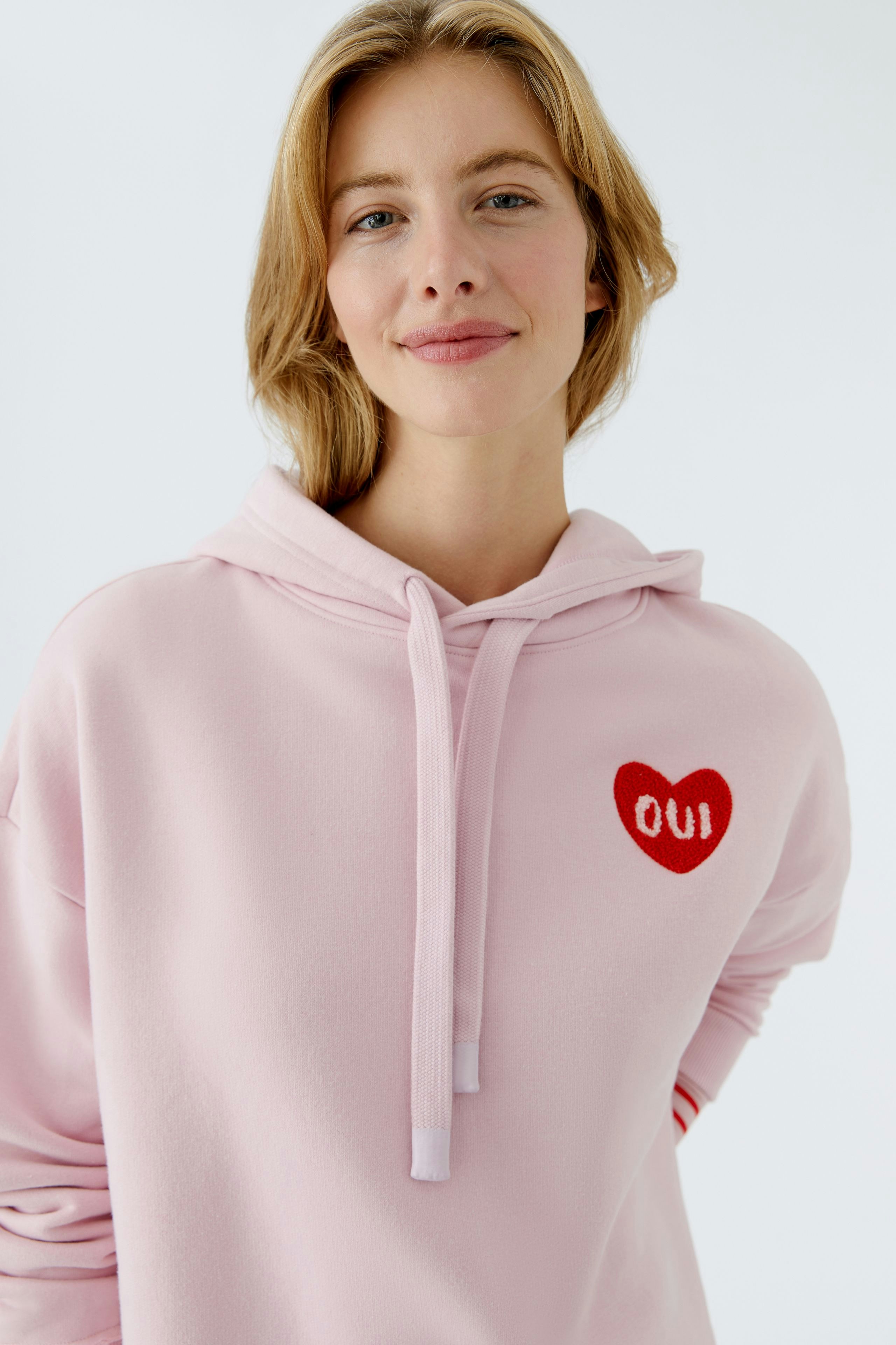 Bequemer Damen-Hoodie: Frau in rosa Kapuzenpullover mit rotem Herz-Logo lächelt freundlich.
