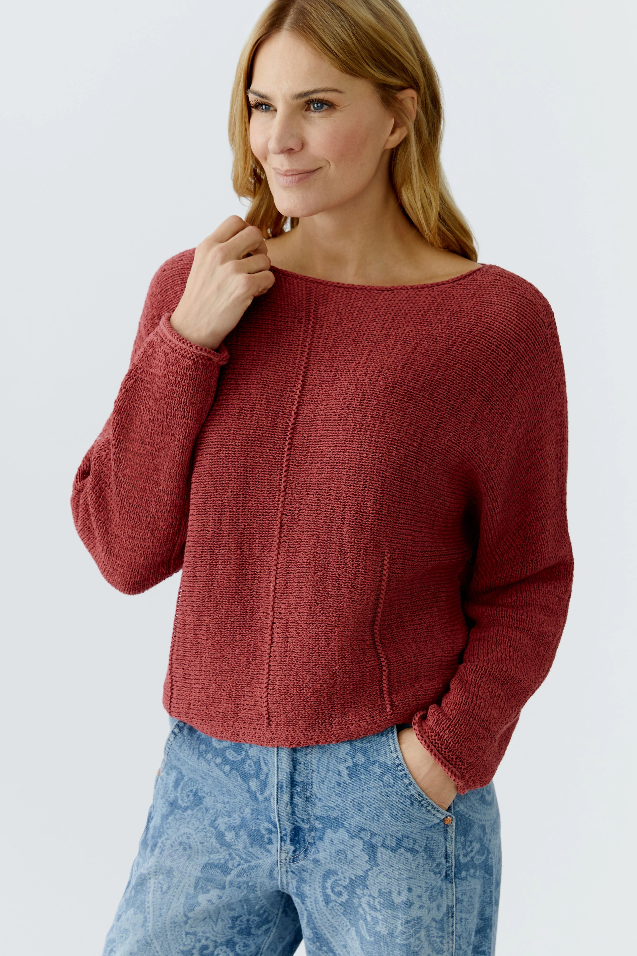 Damen-Pullover: Frau in rotem, lässigem Strickpullover mit weitem Schnitt und Jeans lächelt freundlich.