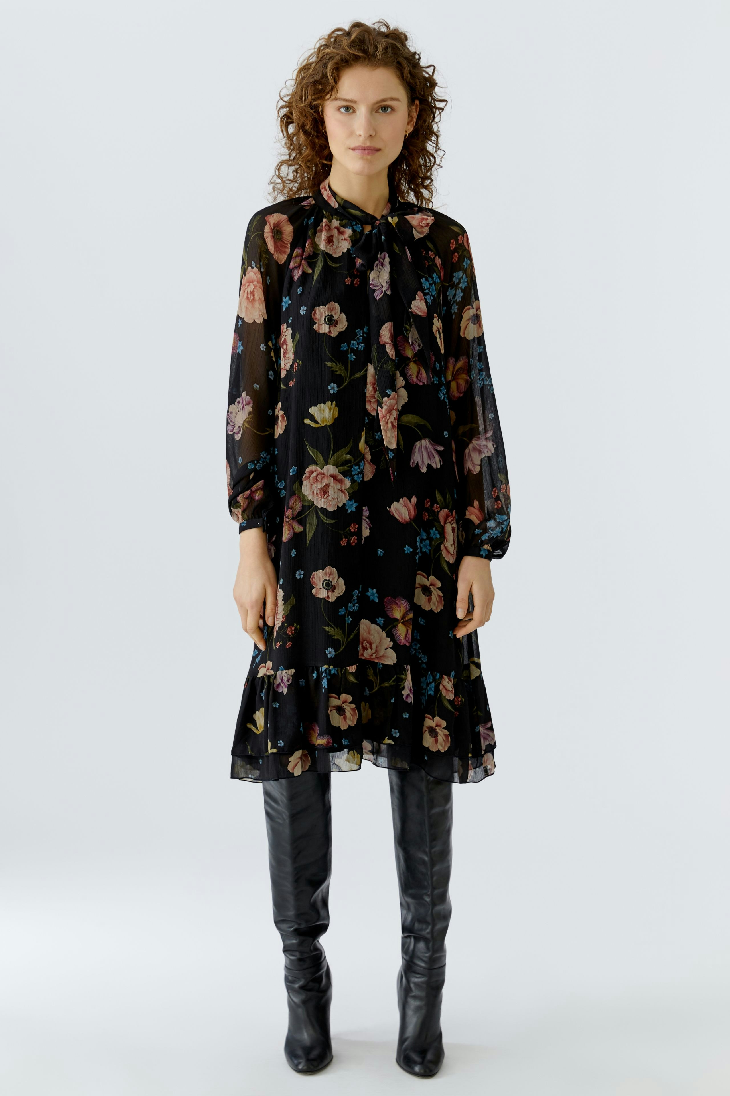 A-Linien-Kleid: Frau in schwarzem, floralem Kleid mit langen Ärmeln und Rüschensaum.