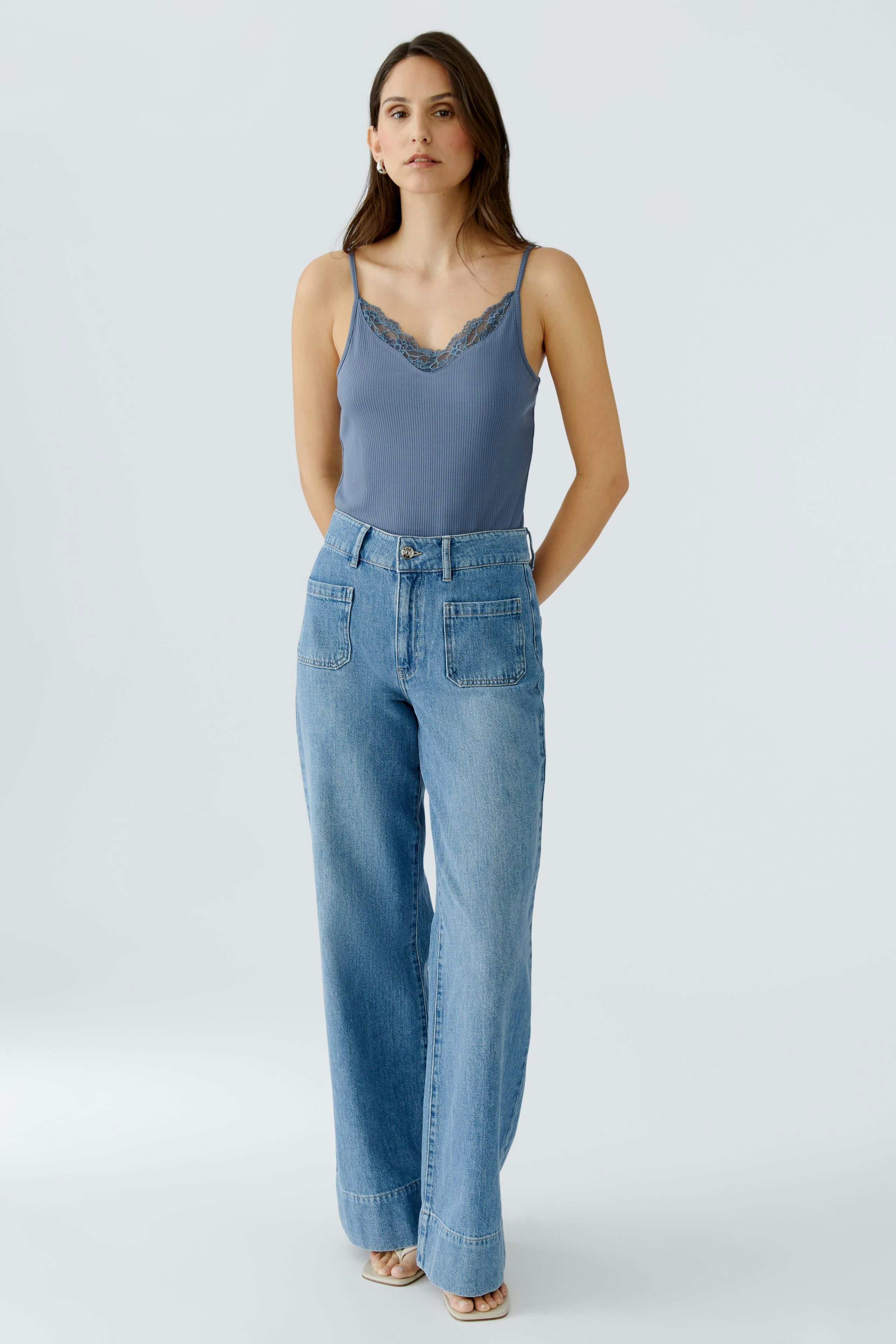 Damen-Top: Junge Frau in einem blauen Top mit Spitzenrand und lässigen Jeans steht entspannt.