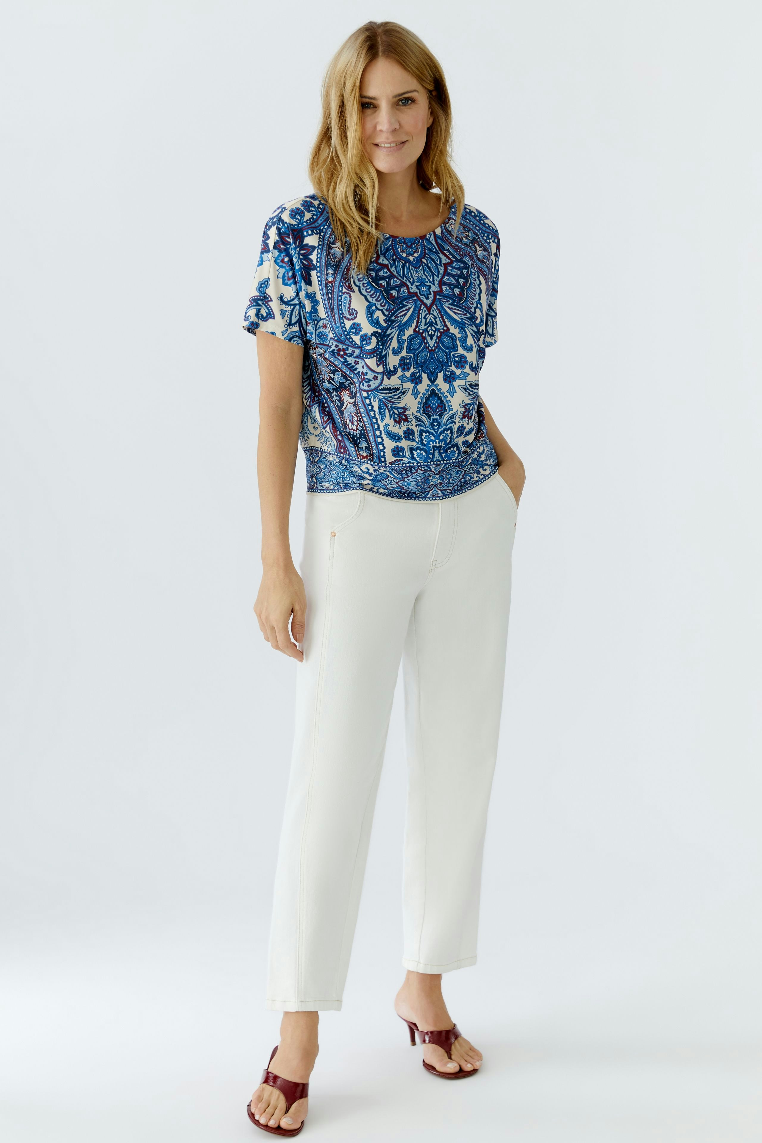 Casual women's blouse shirt: Woman in a blue patterned top paired with white trousers, standing confidently.