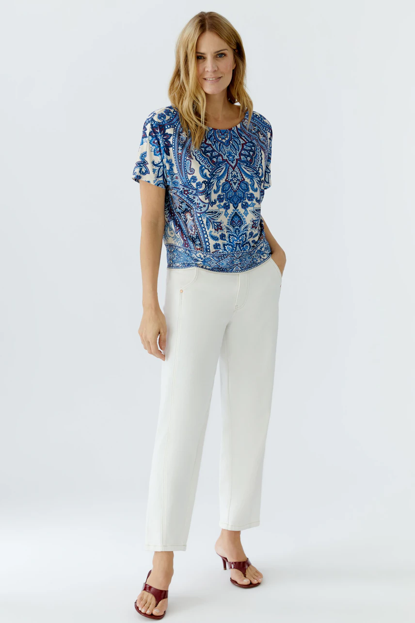 Casual women's blouse shirt: Woman in a blue patterned top paired with white trousers, standing confidently.