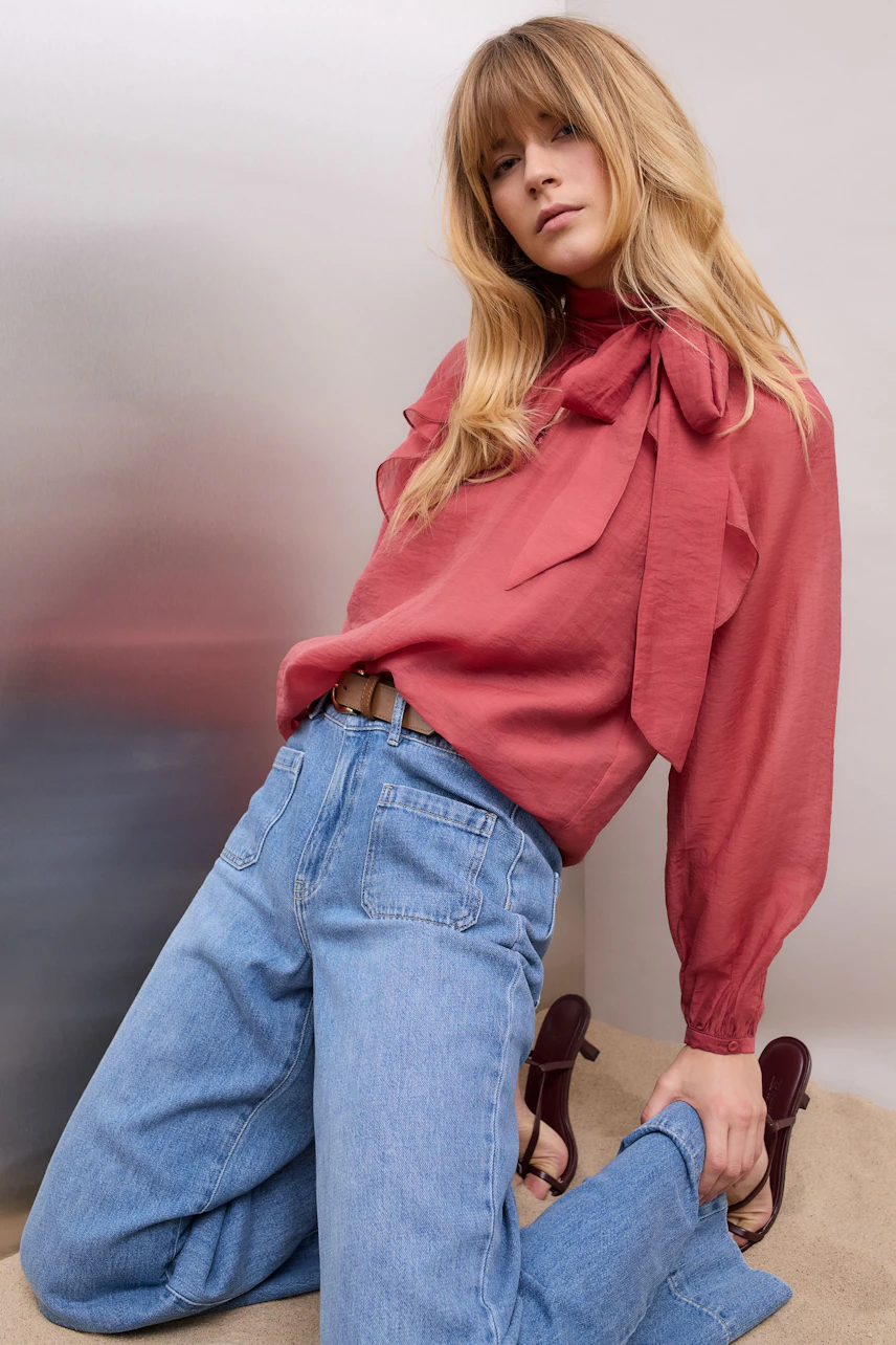 Damen-Bluse: Frau in roter Bluse mit Schleifen-Detail und lässigen Jeans posiert selbstbewusst.