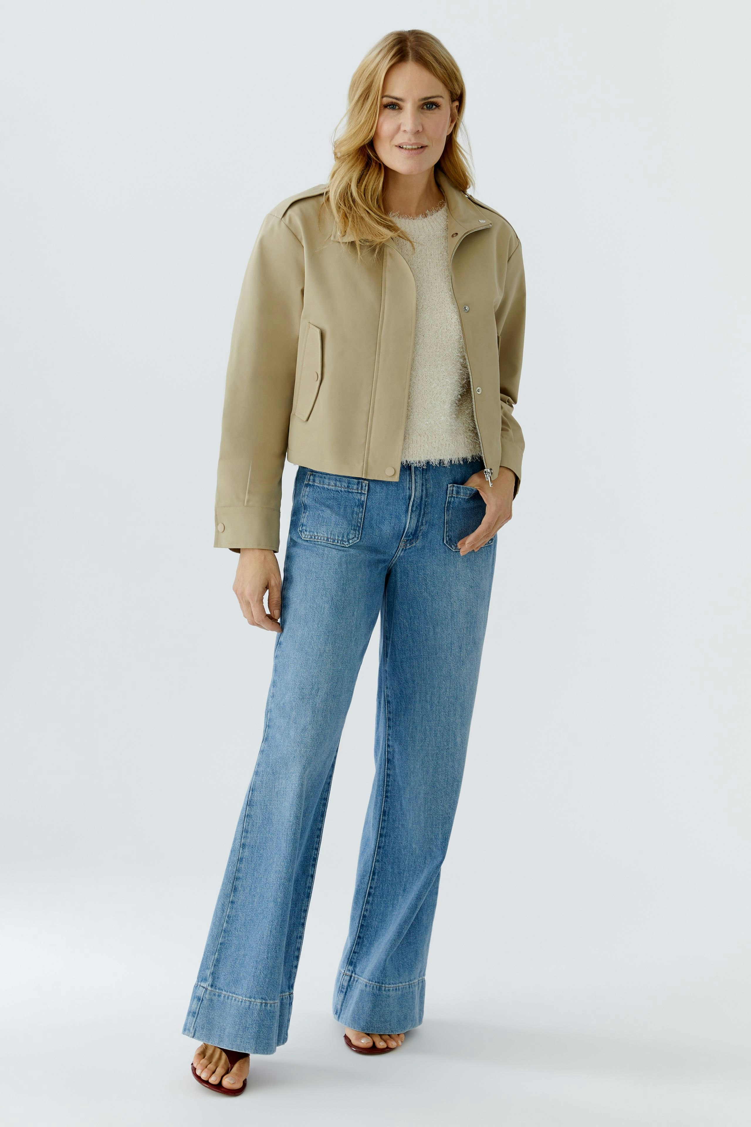 Modern women's blouson: Woman in a beige jacket over a light pullover and flared jeans, smiling confidently.