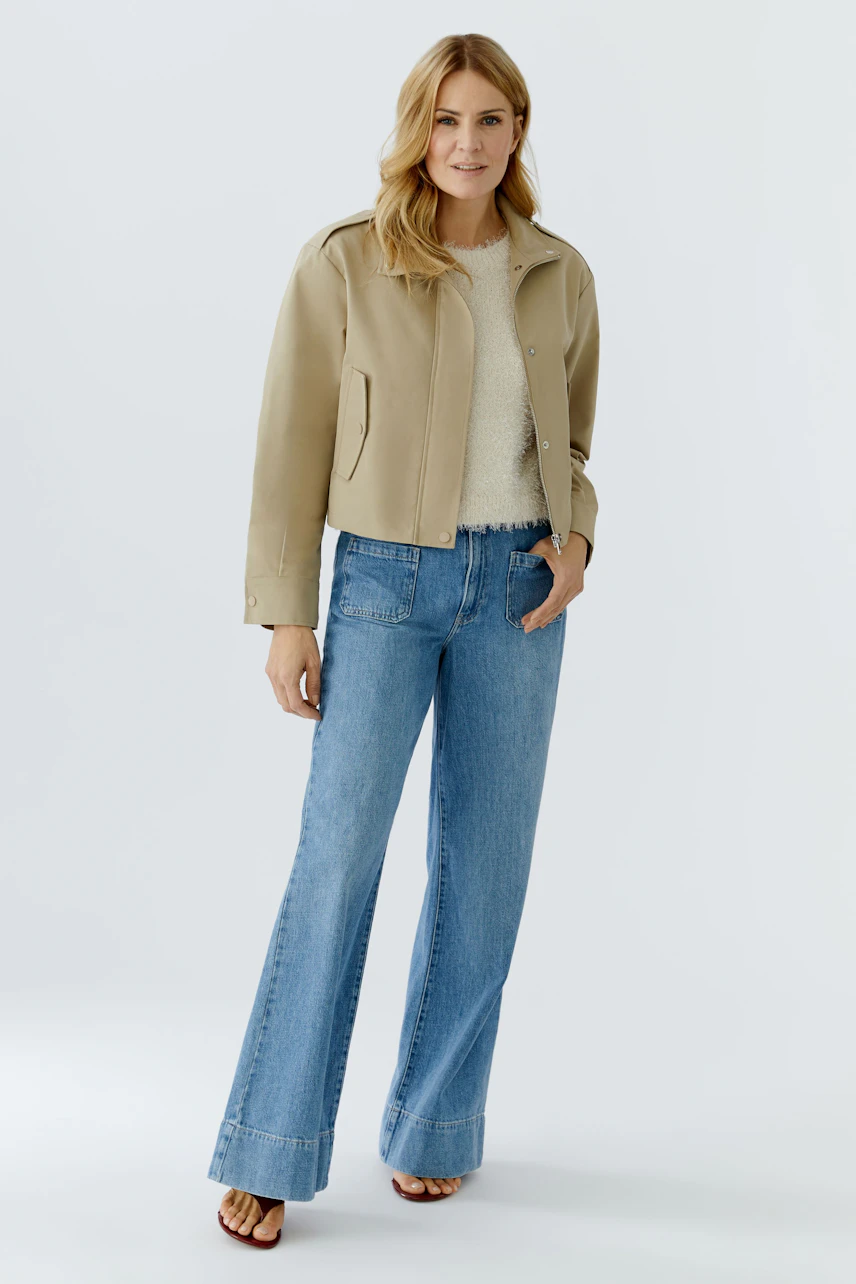 Modern women's blouson: Woman in a beige jacket over a light pullover and flared jeans, smiling confidently.