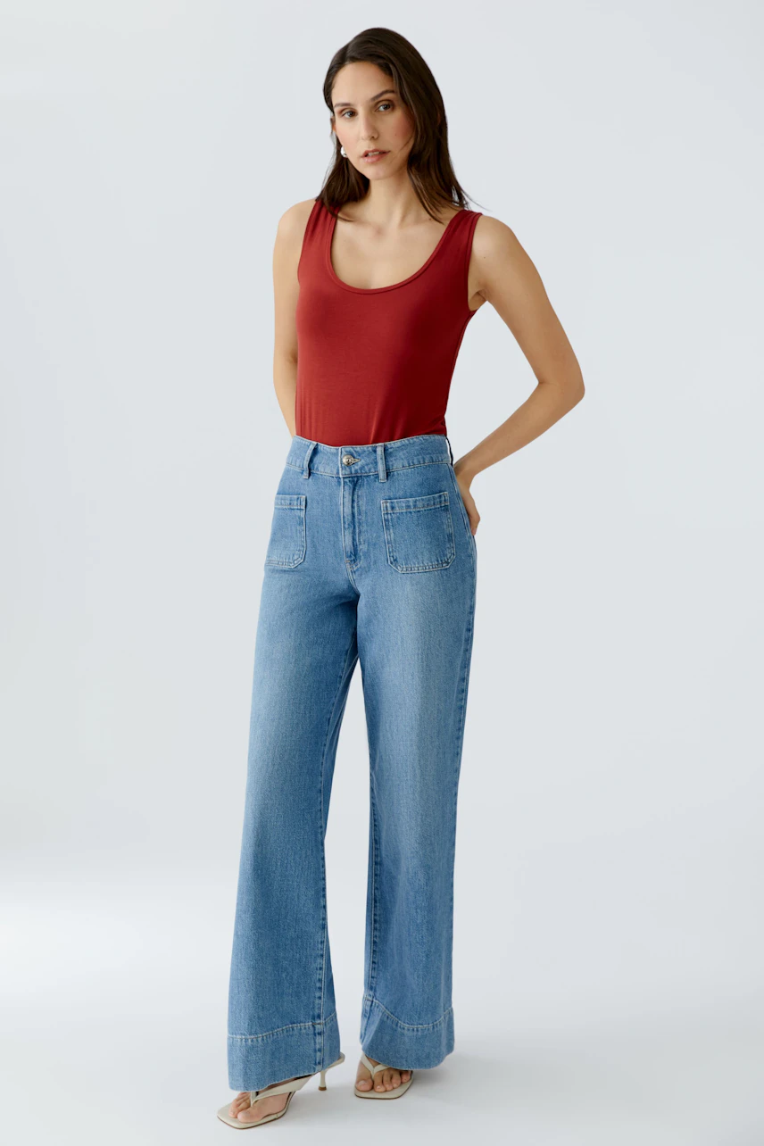 Damen Tank Top: Frau in rotem Tank Top und lässigen Jeans steht entspannt mit den Händen in den Taschen.