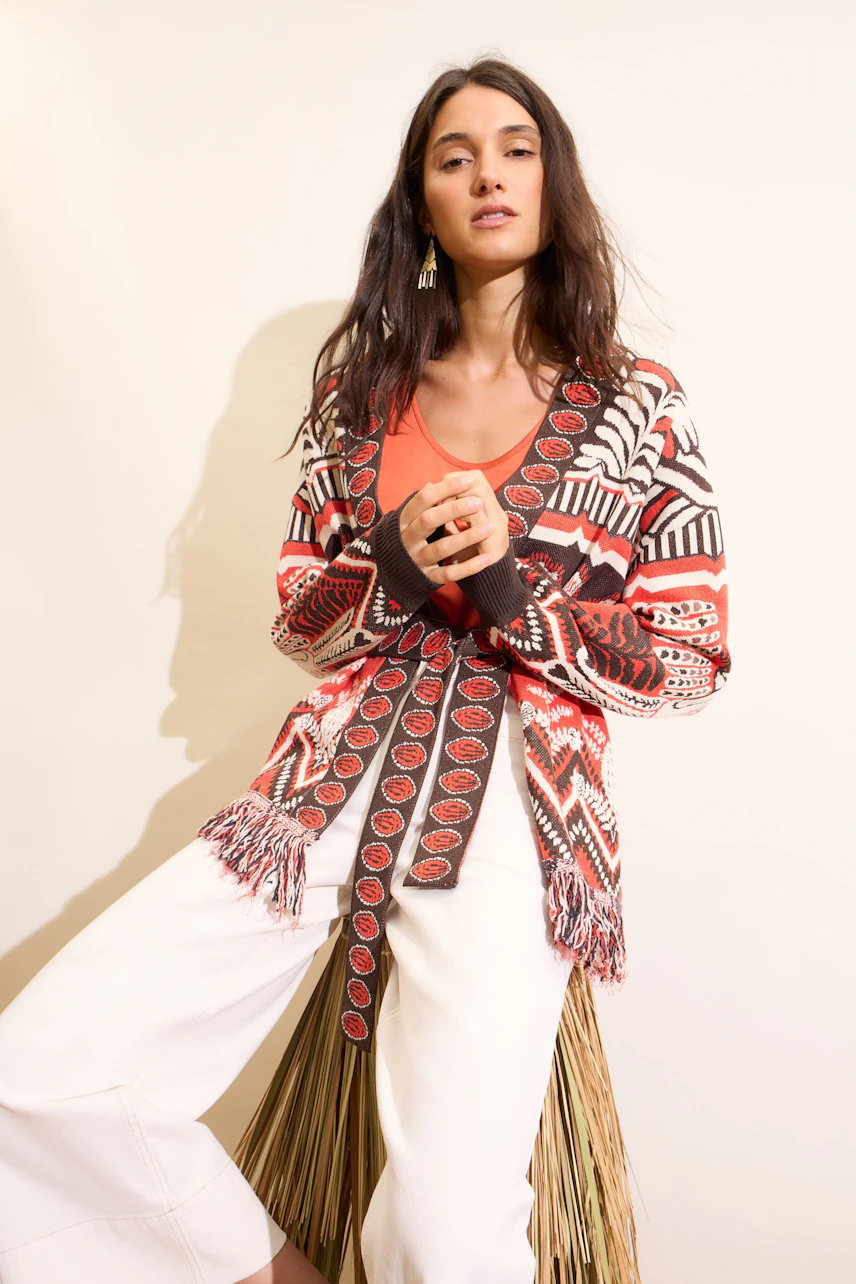 Casual women's top: Woman in a patterned cardigan over a coral top, paired with white pants, seated.