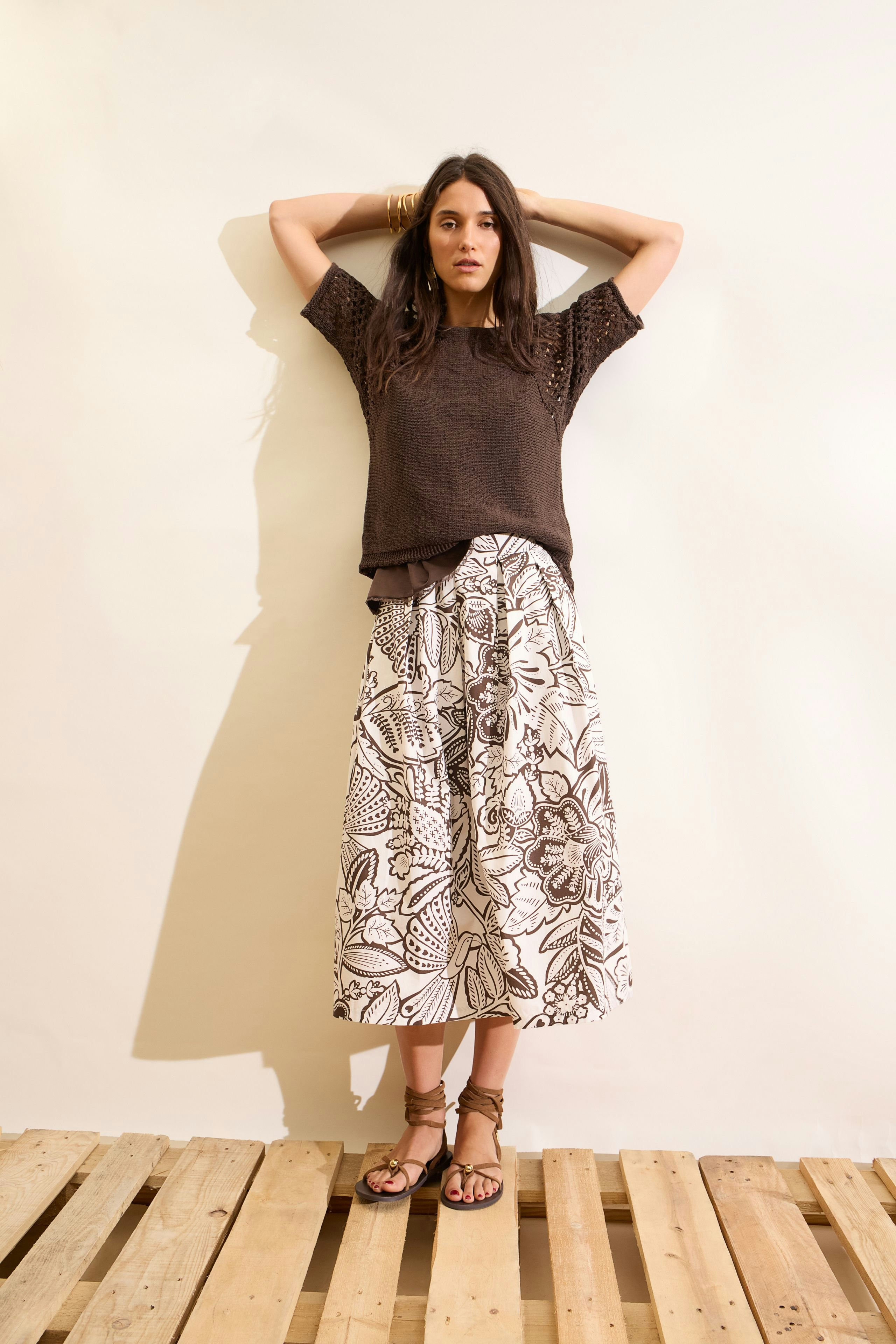 Woman in a floral pleated skirt and brown top, standing confidently with hands on head.