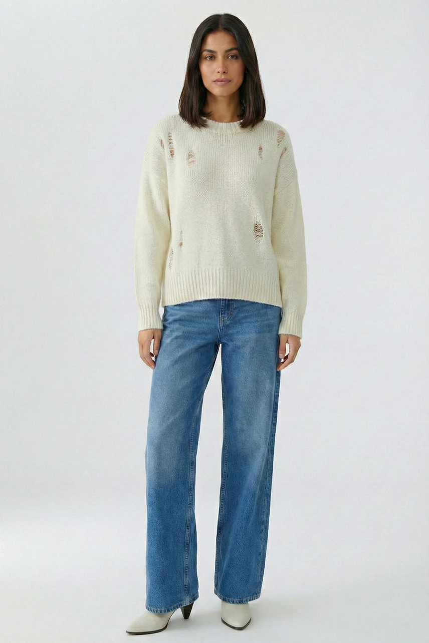 Casual women's pullover: Woman in a distressed cream pullover and blue jeans stands confidently.
