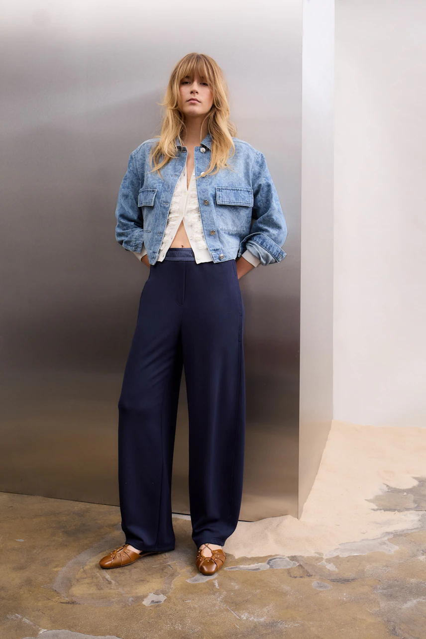 Casual denim jacket: Woman in a blue jeans jacket, white top, and navy pants stands confidently.