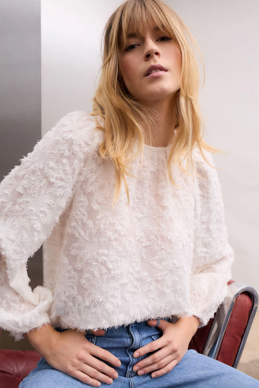 Frau in weißer Bluse mit Strukturmuster, lässig kombiniert mit blauer Jeans, sitzt entspannt.