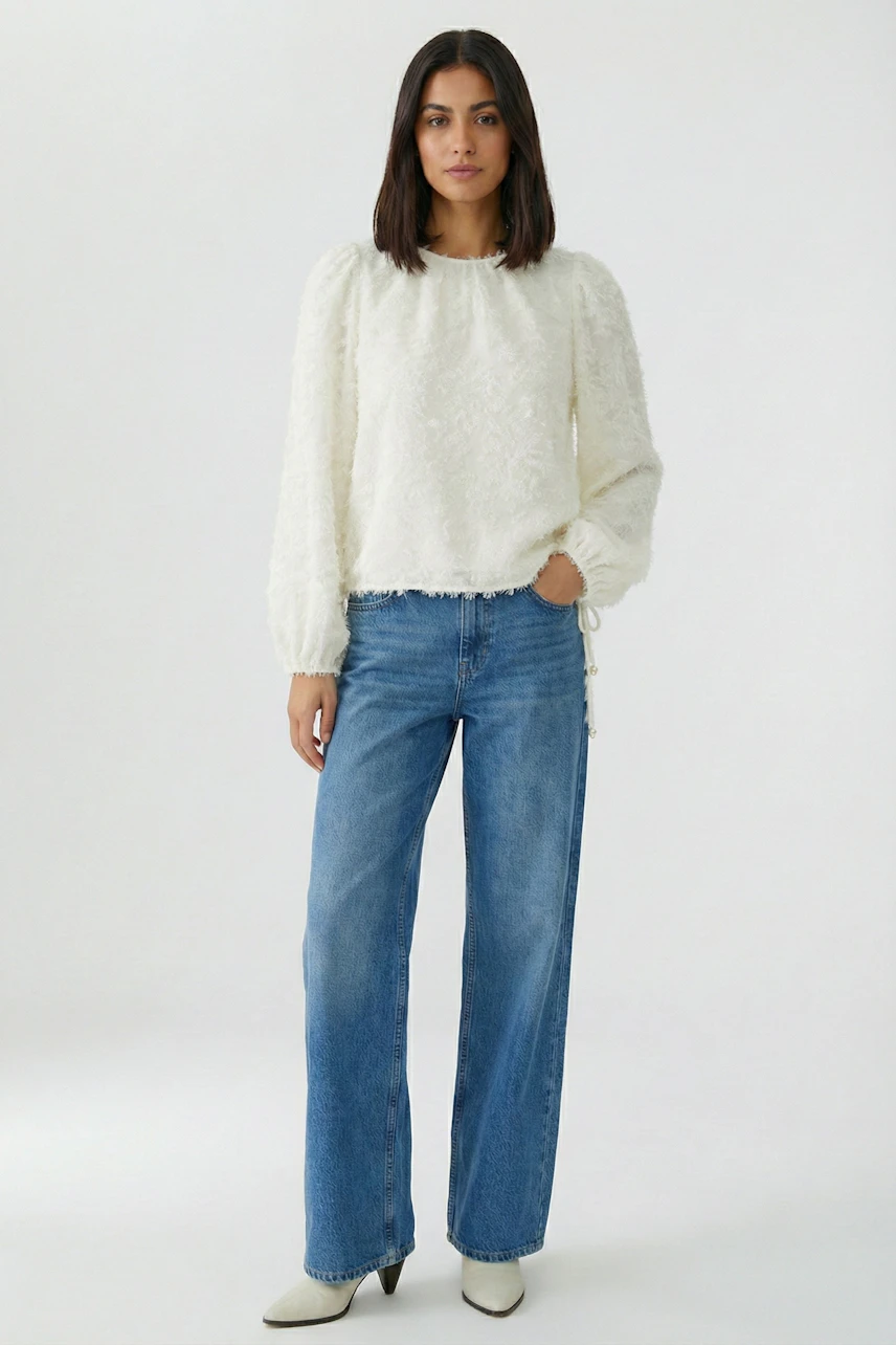 Frau in weißer Bluse und blauer Jeans. Die Bluse hat lange Ärmel und eine strukturierte Oberfläche.