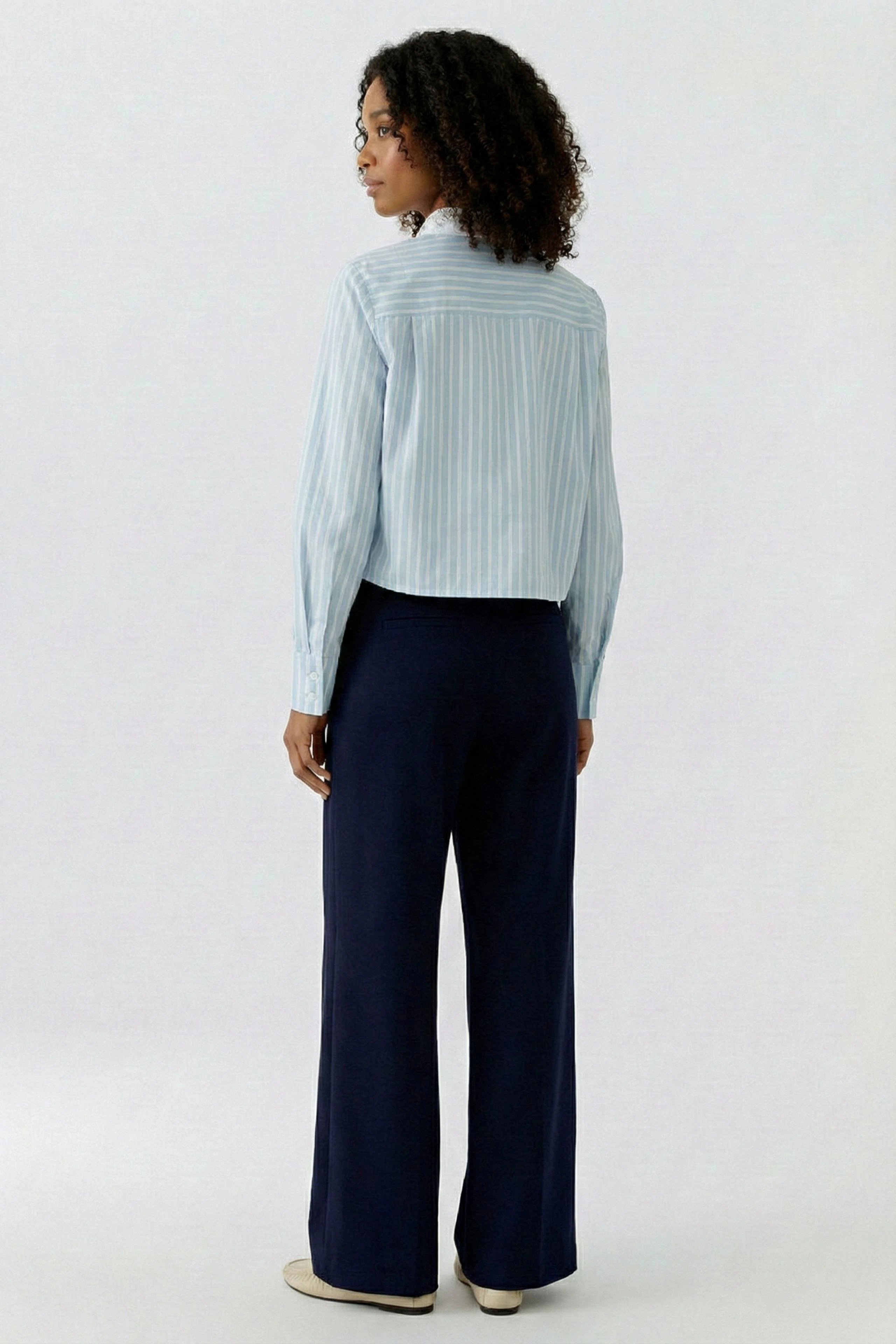 Damen-Bluse: Frau in gestreifter Bluse und dunkler Hose, von hinten gesehen, mit lockigem Haar.