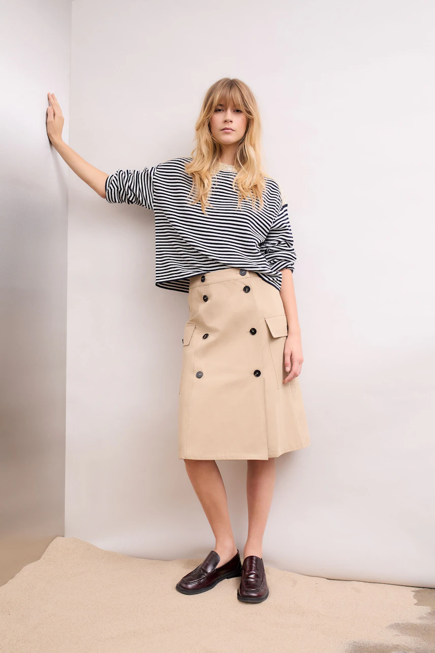A-Linien-Rock: Frau im gestreiften Pullover und beigem Rock mit Knöpfen lehnt lässig an der Wand.
