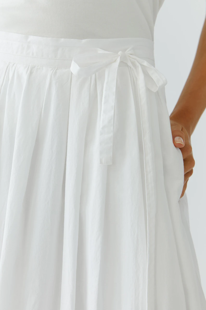 White pleated skirt: Woman wearing a flowy white skirt with a tied waistband and pockets.