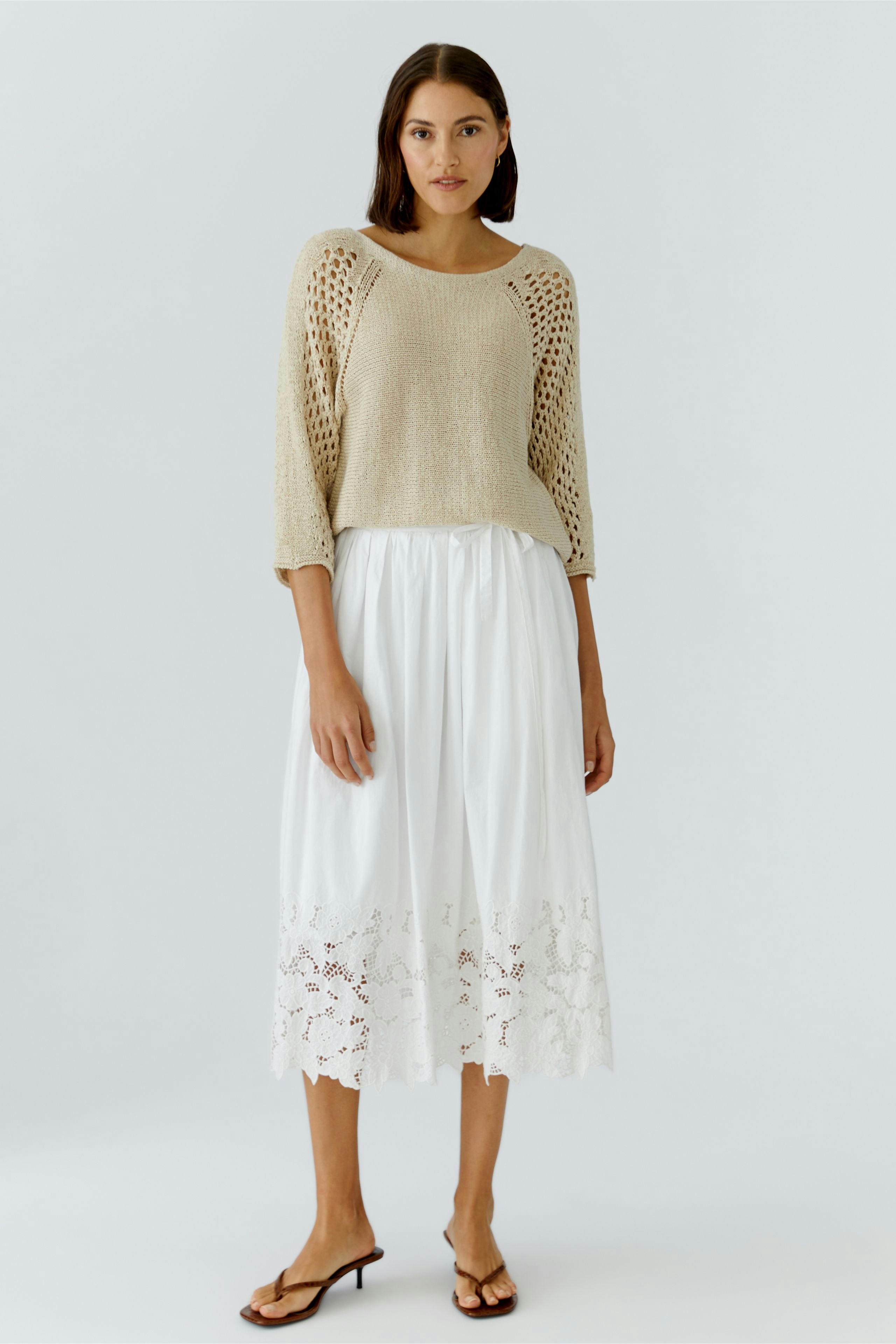 A woman in a light knit pullover and a white pleated skirt with lace detailing stands confidently.
