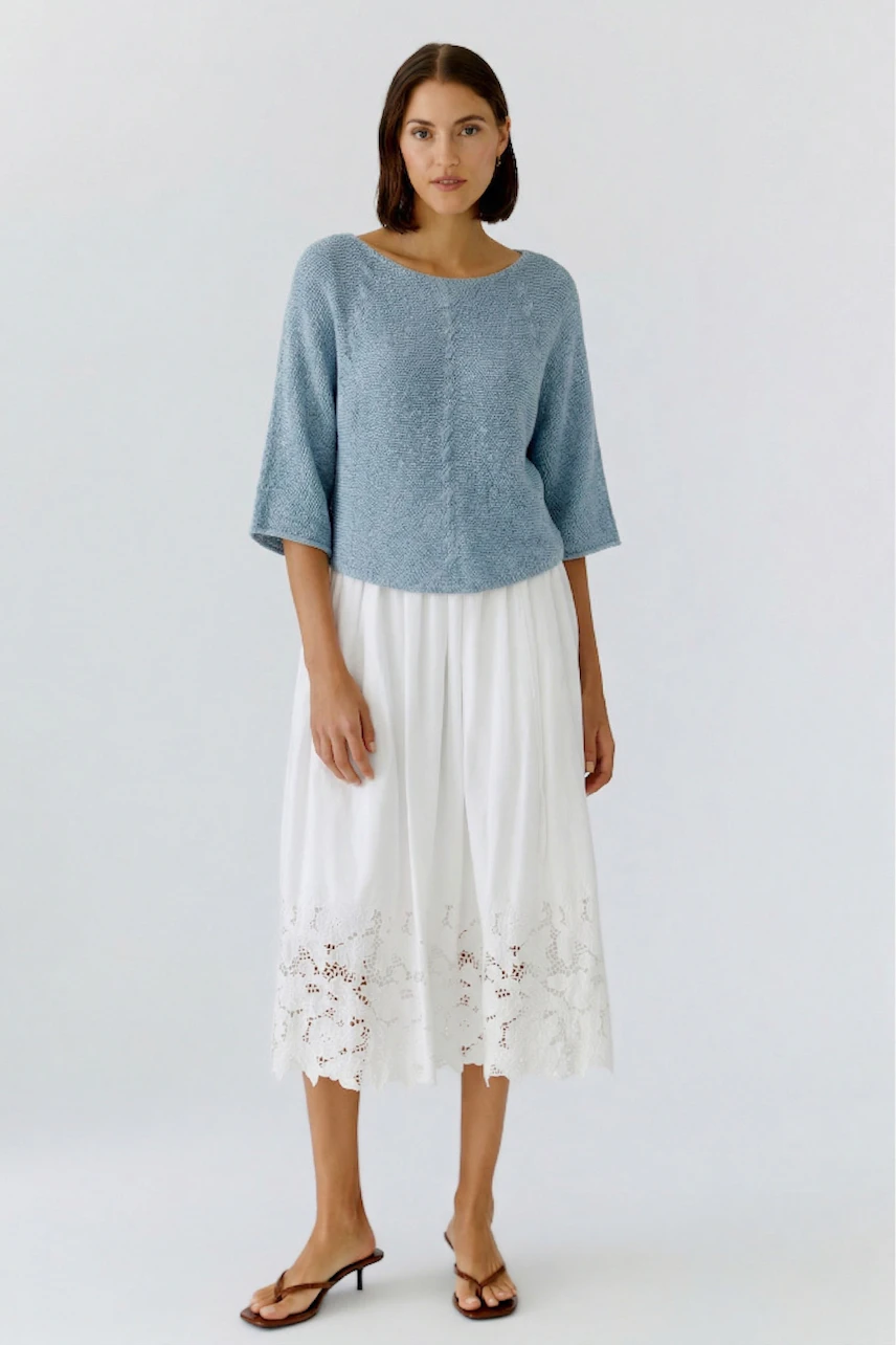 A woman in a light knit pullover and a white pleated skirt with lace detailing stands confidently.