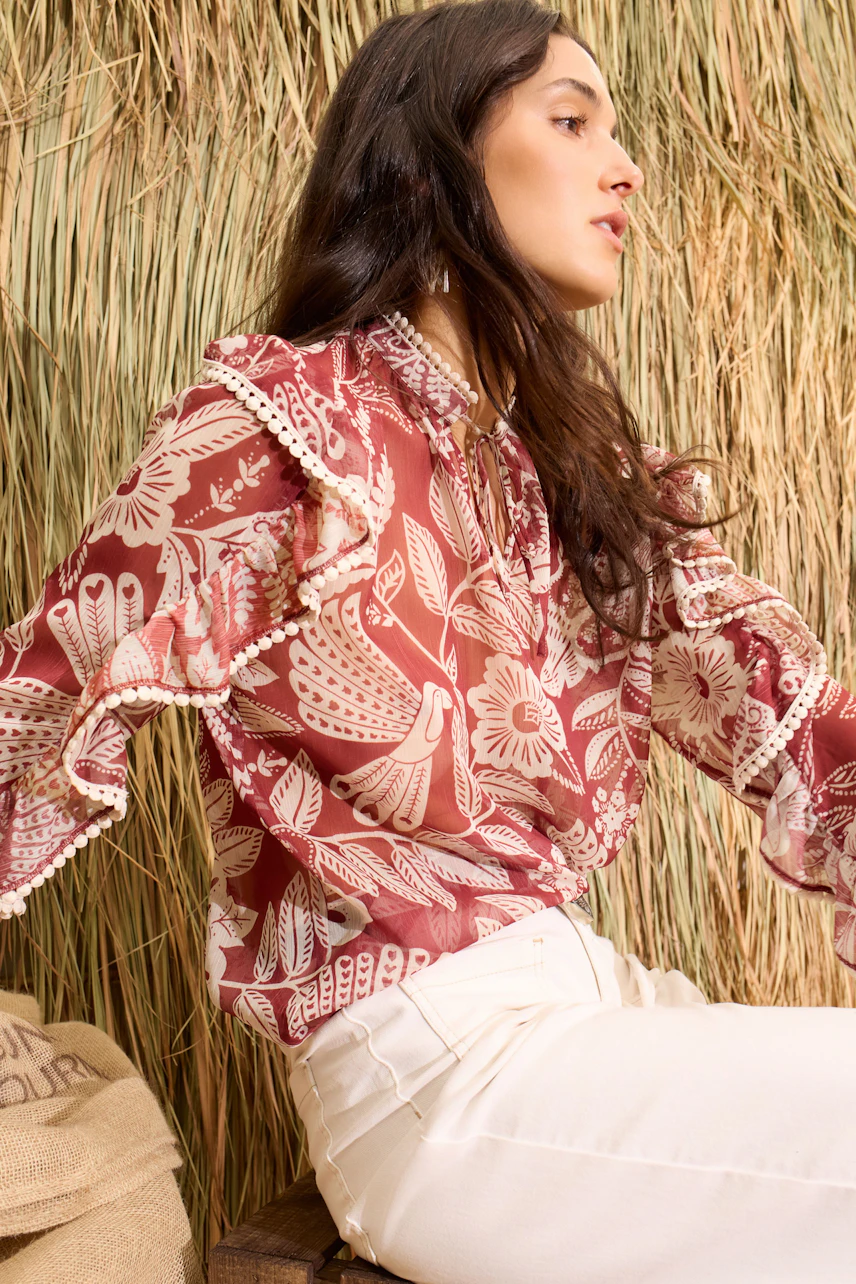 Frau in gemusterter Bluse: Rote Bluse mit floralen Motiven, kombiniert mit weißen Hosen, vor natürlichem Hintergrund.