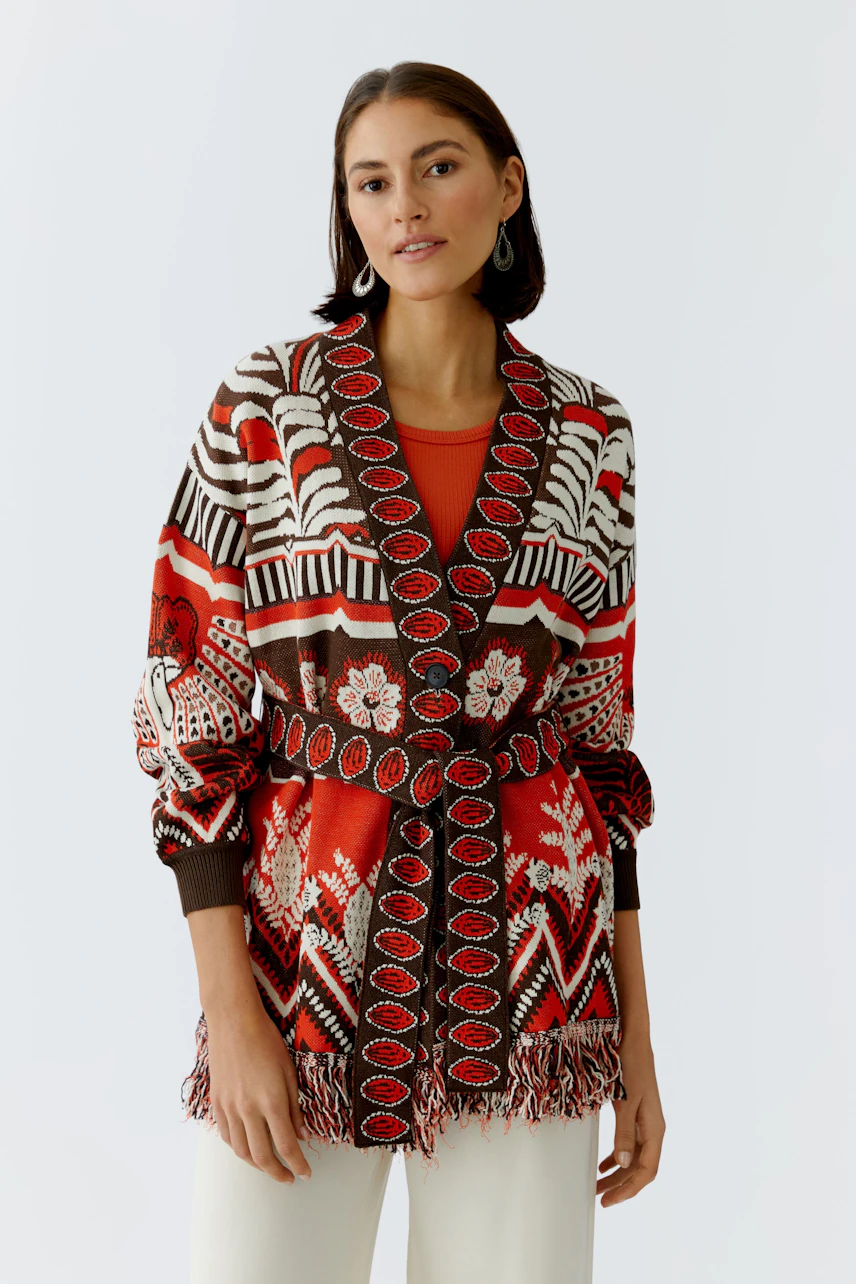 Colorful women's cardigan with a patterned design and fringe details, worn over a red top.