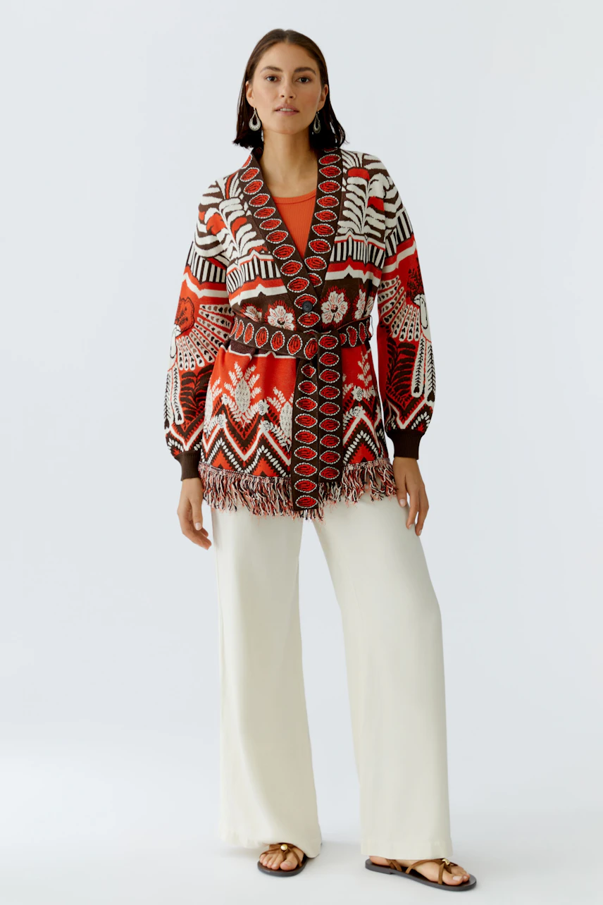 Colorful women's cardigan with a bold pattern and fringe details, styled with wide pants.