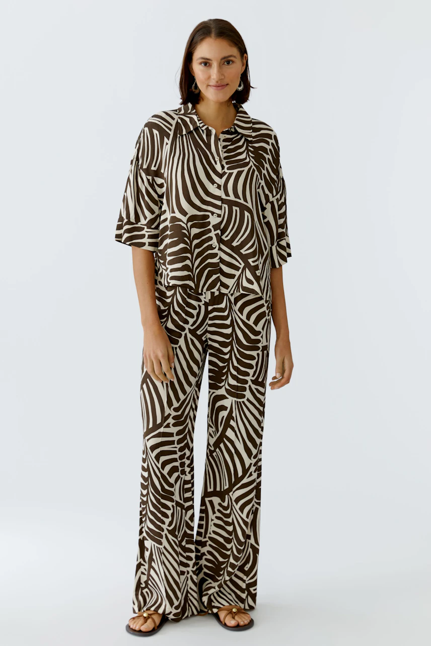 Casual women's blouse: Woman in a brown and cream patterned blouse and matching pants, standing confidently.