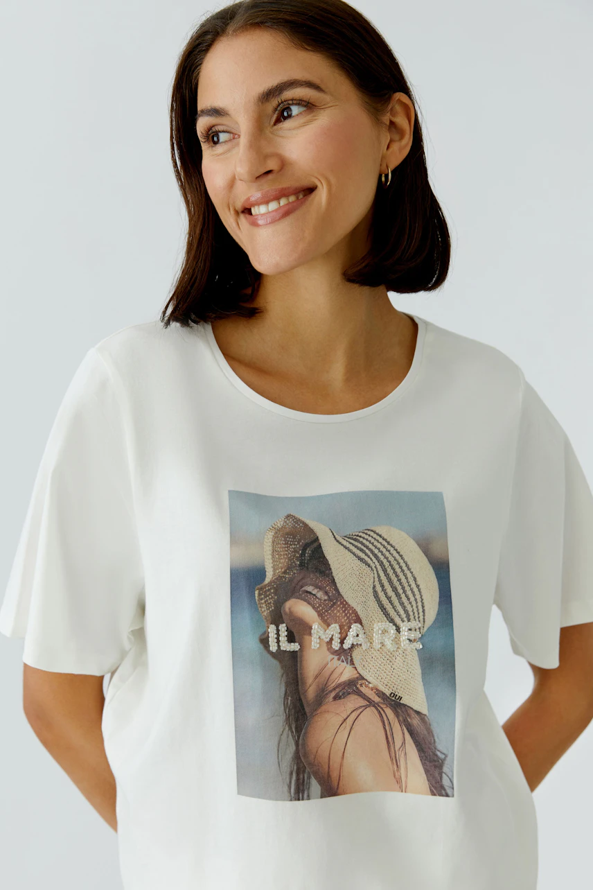Casual women's T-shirt: Woman smiling in a white tee featuring a beach hat graphic.