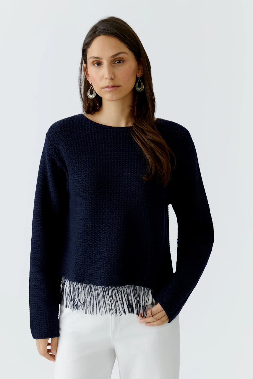 Casual women's pullover: Young woman in a navy pullover with fringe detail, paired with white pants.