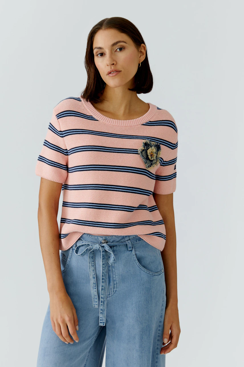 Casual women's pullover: Young woman in a pink striped pullover with a lion patch, paired with light jeans.