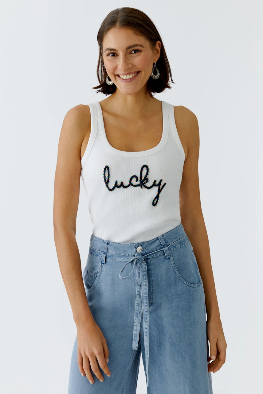 Casual women's tank top: A woman in a white tank top with 'lucky' print, paired with light blue wide-leg pants.