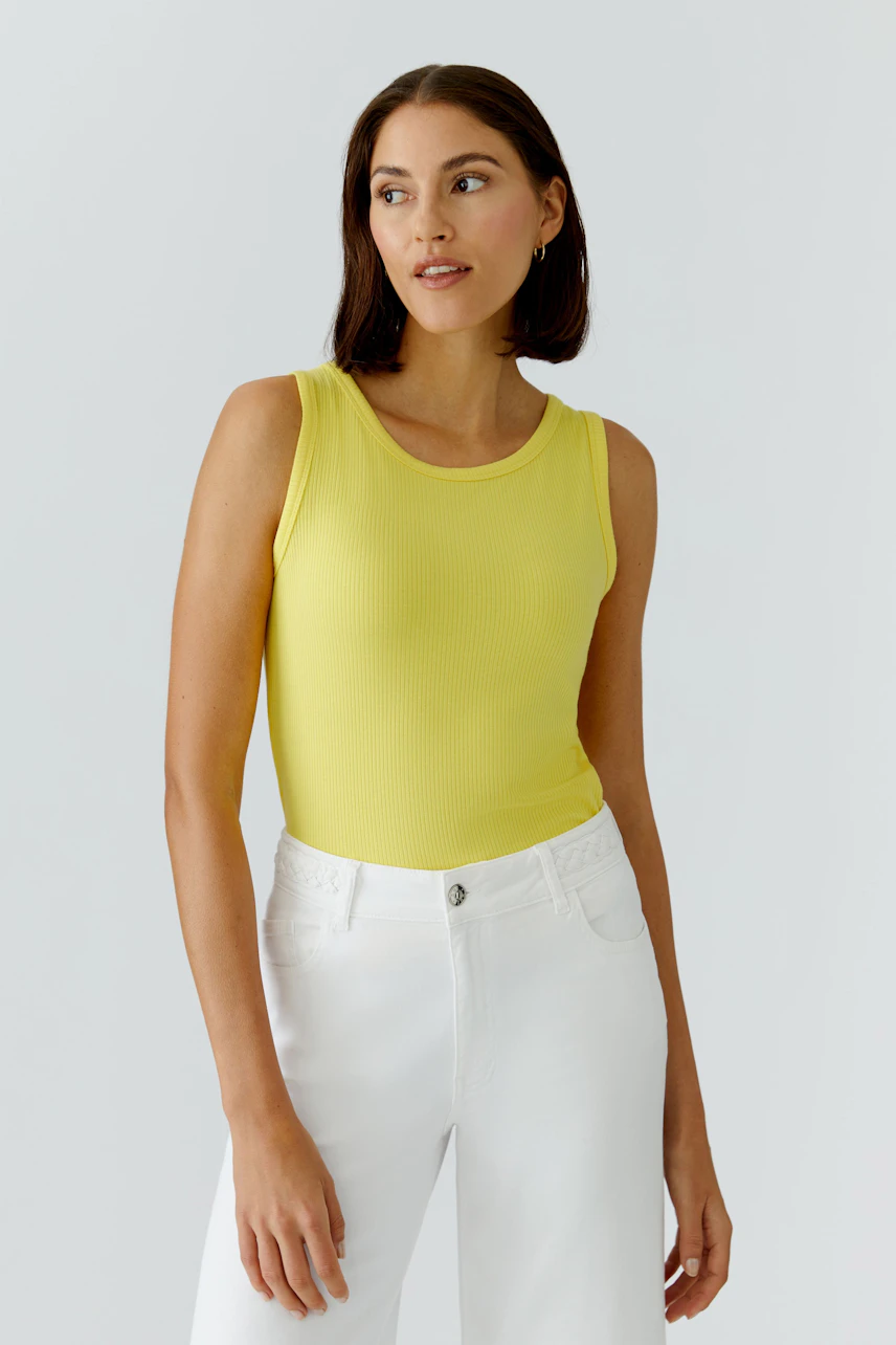 Casual women's top: Woman in a yellow sleeveless top and white pants, looking to the side.