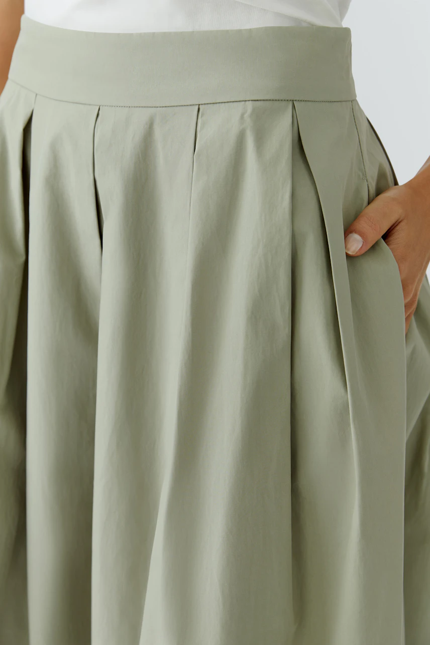 A-line skirt: Woman in a light green A-line skirt with pleats, hand in pocket, wearing a white top.