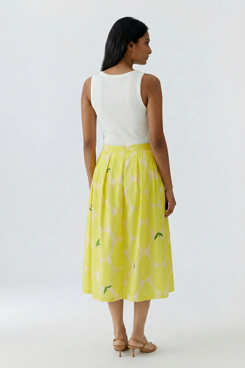 Woman in a yellow pleated skirt and white tank top, standing confidently. Casual and stylish look.