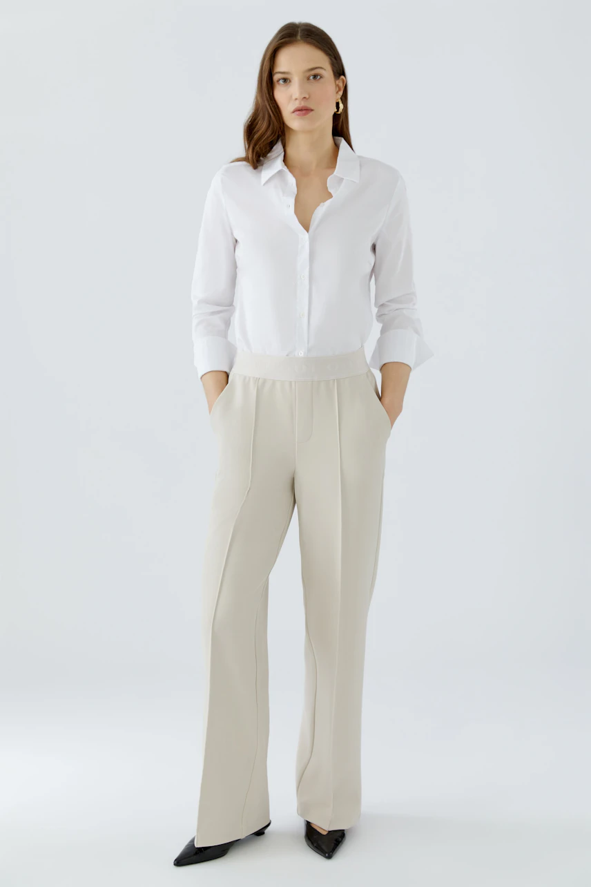Chic women's trousers: A woman in beige trousers with a relaxed fit and a white shirt, hands in pockets.