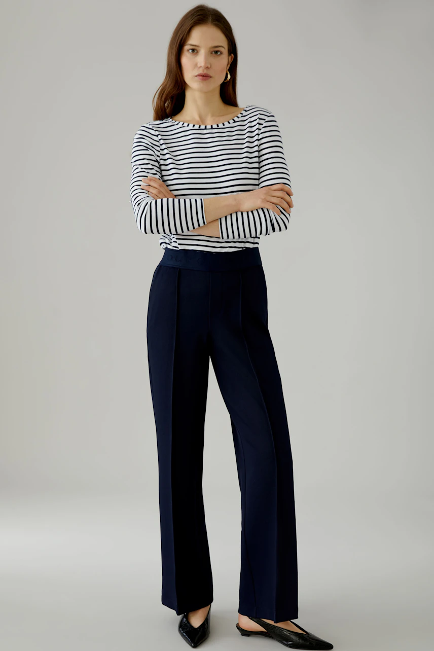 Woman in navy trousers and white shirt, standing confidently. The trousers are high-waisted and tailored.