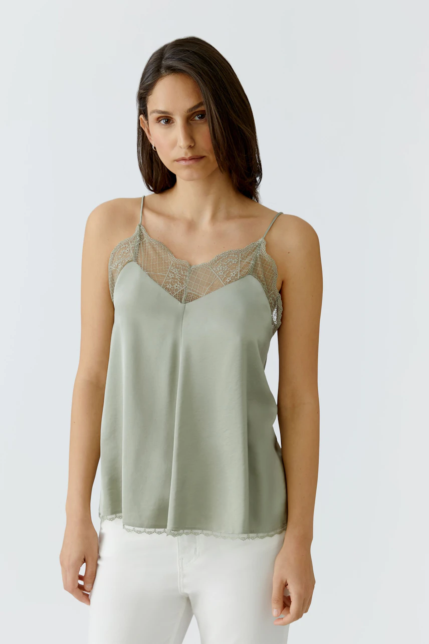 Casual women's top: Woman in a light green top with lace details, paired with white pants, looking relaxed.